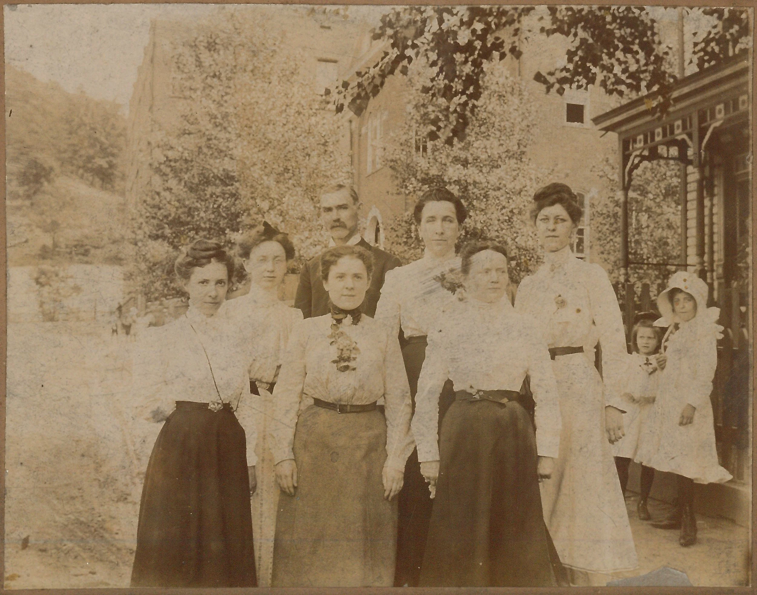 A vintage sepia photograph of Beulah Cockayne and fellow teachers. The women are dressed in long skirts and blouses, some with corsages, and the man is in a suit. Two children are in the background.