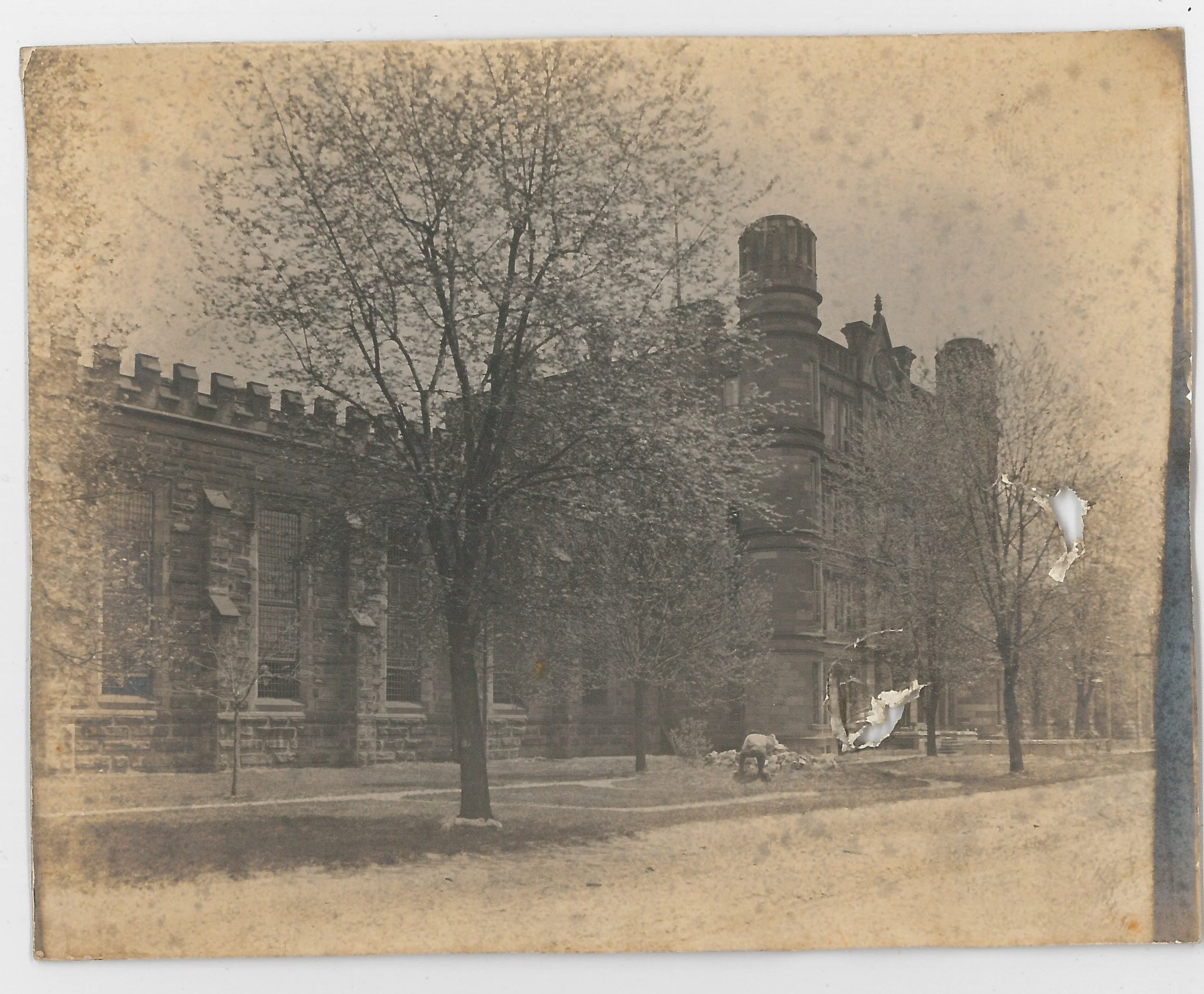 An old black-and-white photograph of a historic building with a tower, surrounded by leafless trees and a grassy area.