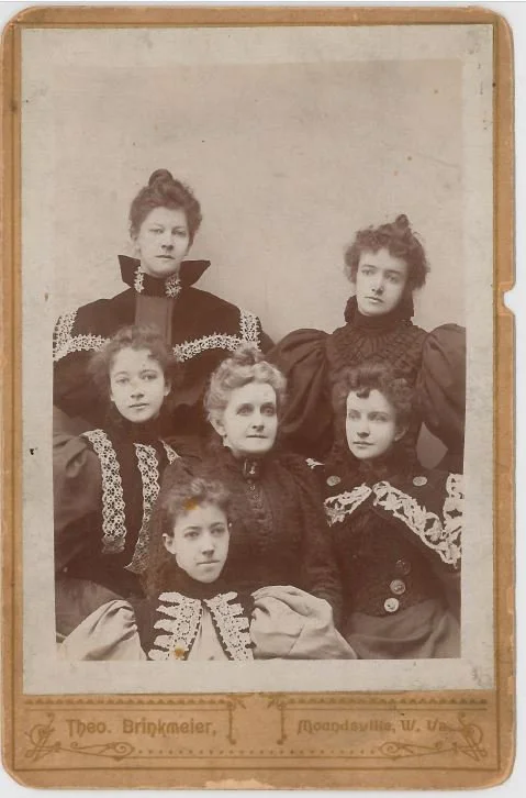 Historic black-and-white portrait of six women dressed in Victorian-style clothing, arranged in two rows, with the two women in front seated and the four women standing behind them. The photo has a labeled border that includes the photographer's name, Theo Brinkmeier, and location, Moundsville, West Virginia.