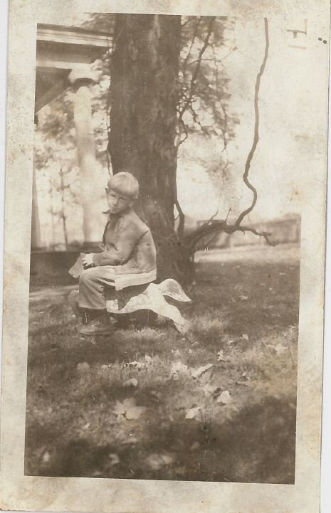 A young Sam Cockayne sitting on a toy horse near a tree outdoors, with bare branches and a grassy area with fallen leaves.