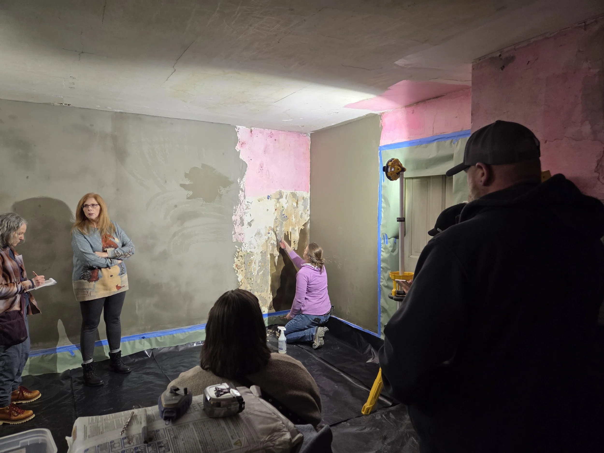 Group of people participating in a wall renovation project inside a room, with some individuals taking notes and others actively working on the wall.