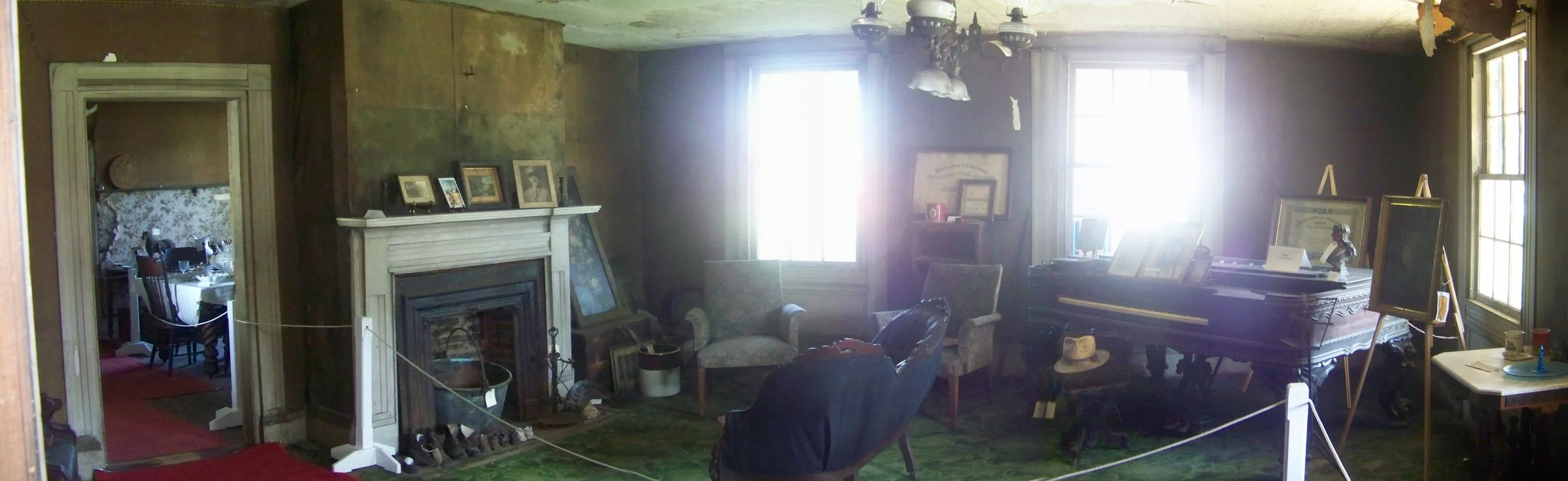 A cluttered, vintage living room with old furniture, a fireplace with framed photos on the mantle, a piano, and large windows letting in bright sunlight.