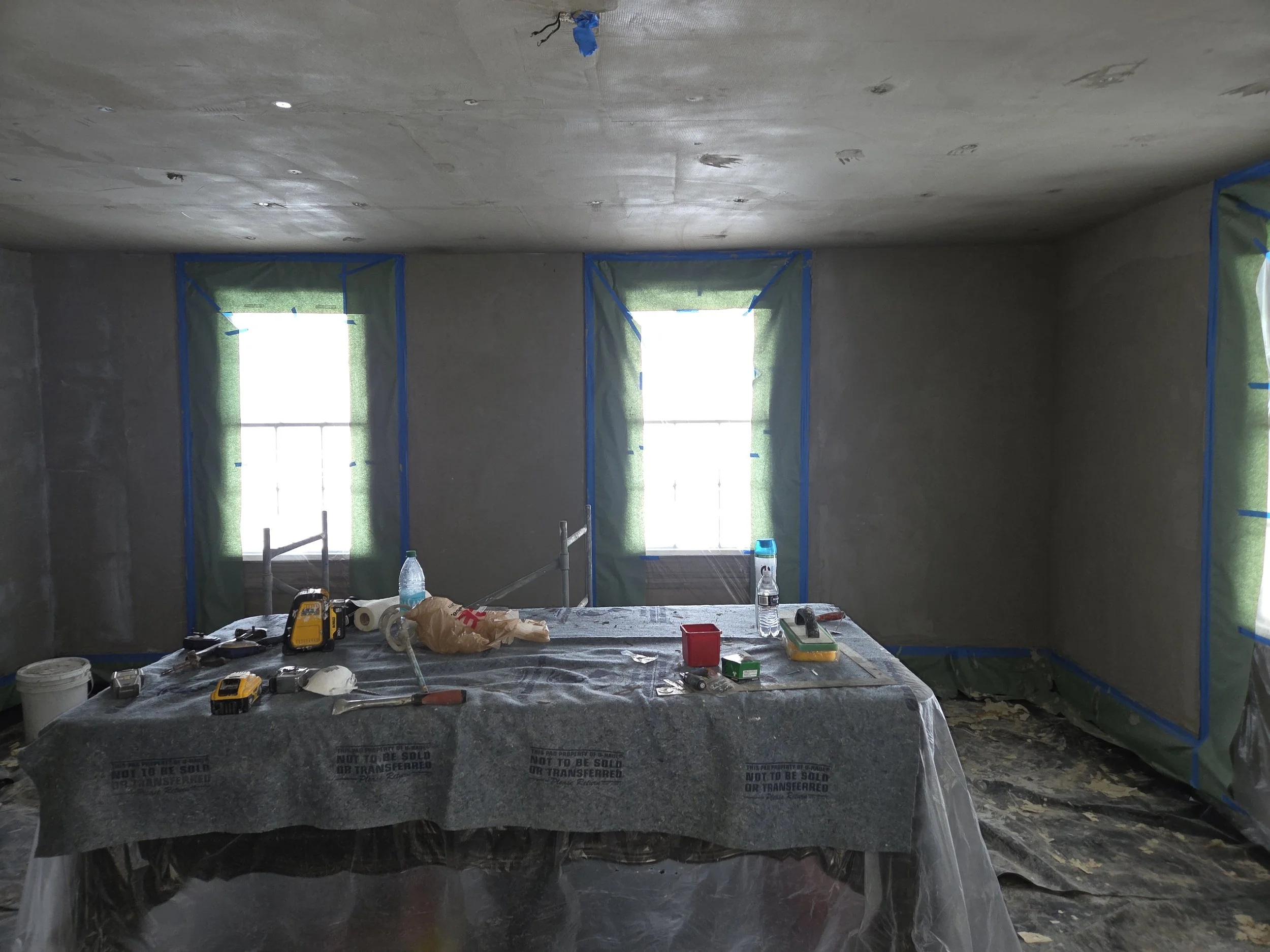 Interior of a room under renovation with three windows covered in green fabric, plastic-covered table in the center with tools and supplies, and a concrete floor.