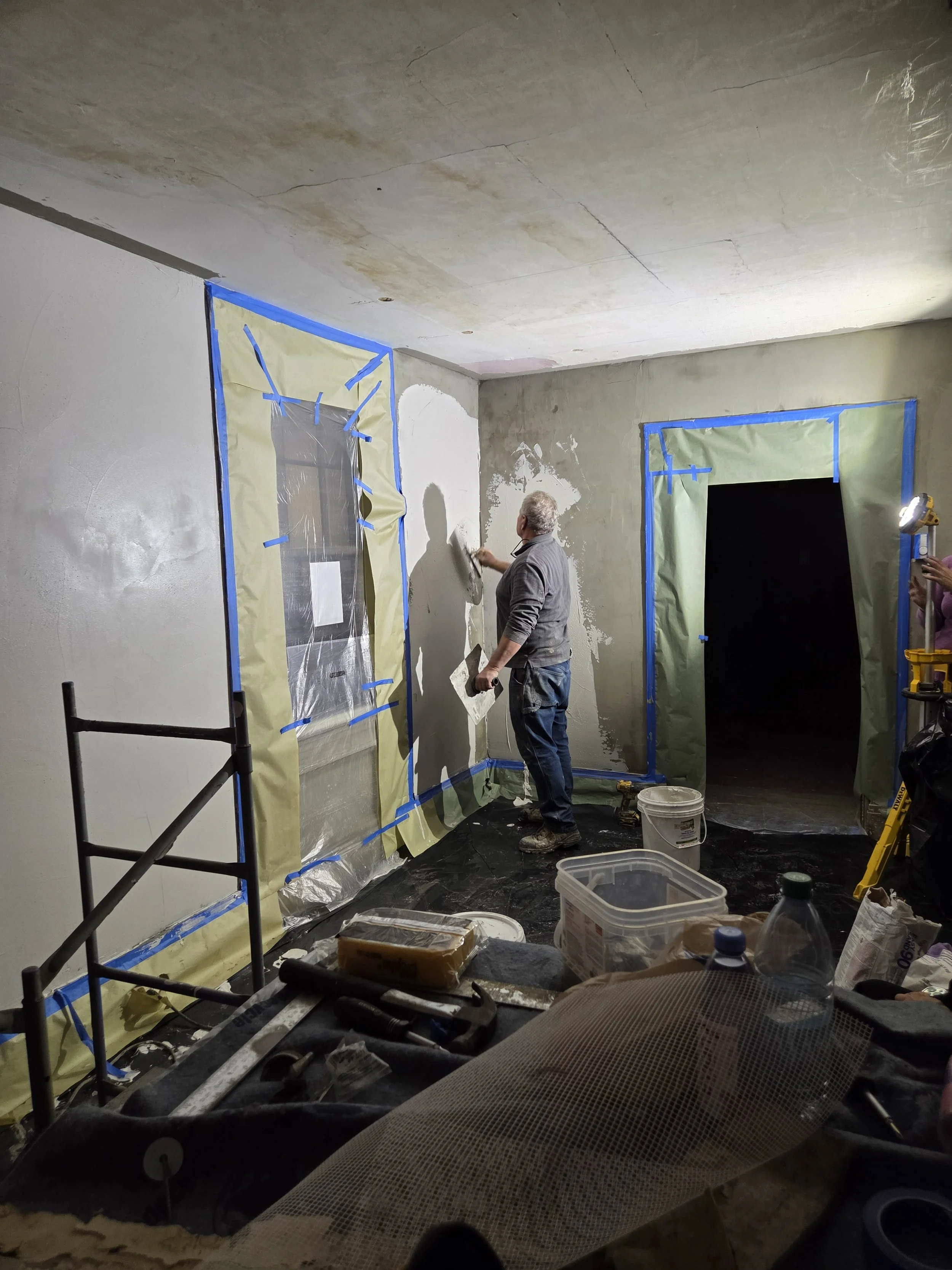 A man is applying plaster to a wall in a room under renovation, with windows covered in protective paper and painter's tape, and construction tools and supplies scattered on the floor.