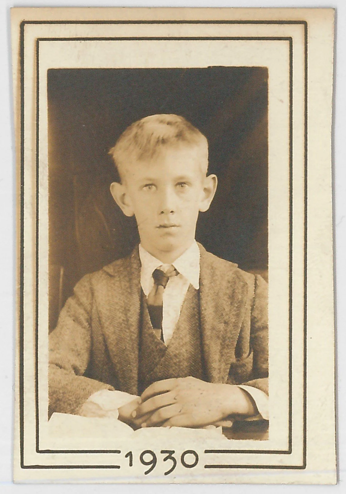 A black and white vintage portrait photograph of a young boy, around 10 years old, dressed in formal attire with a blazer, shirt, tie, and vest, sitting with hands clasped, from the year 1930.