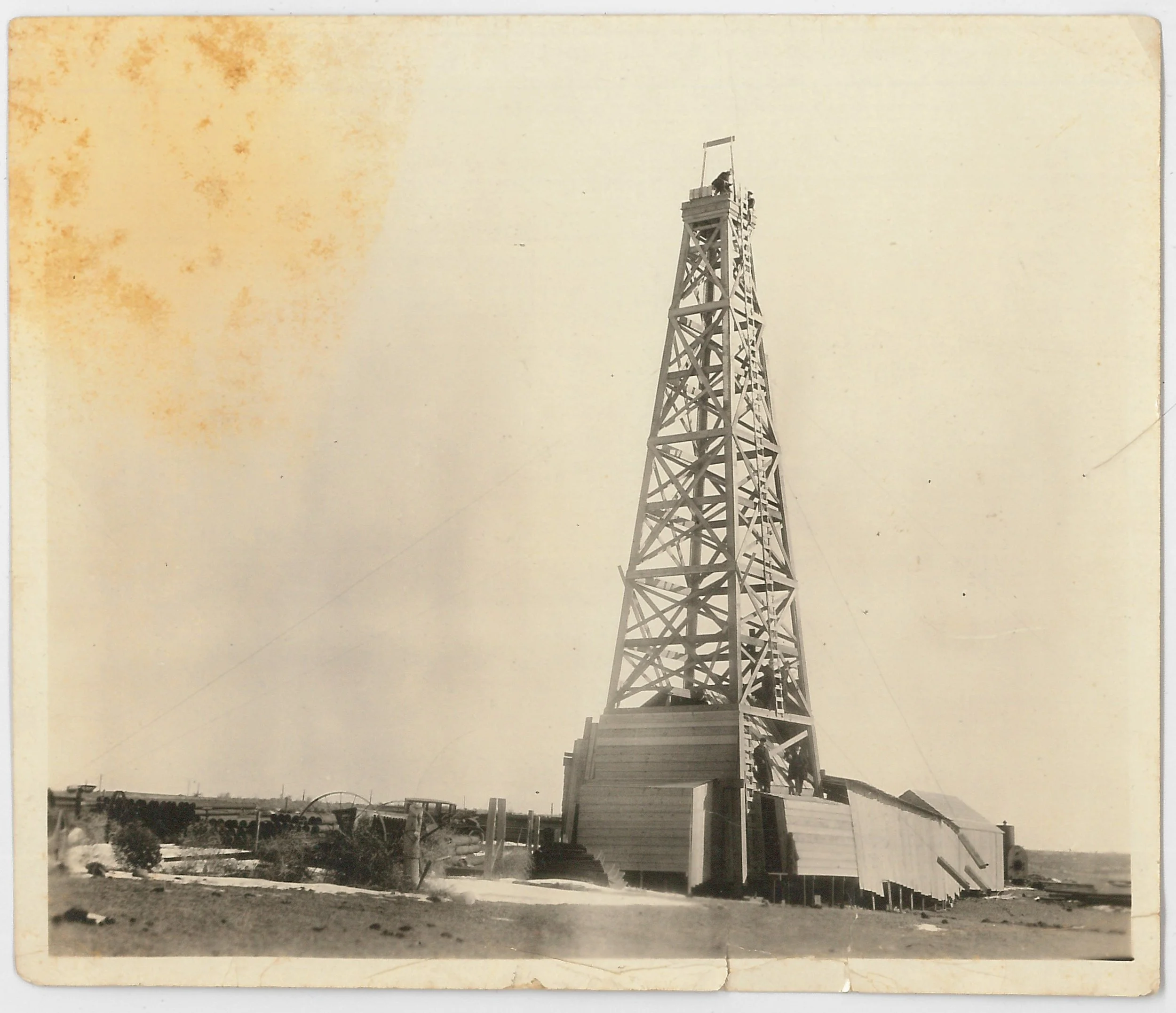 An old black-and-white photograph of a tall wooden oil derrick with a small platform and ladder at the top, situated in an open landscape with some buildings and trees in the distance.