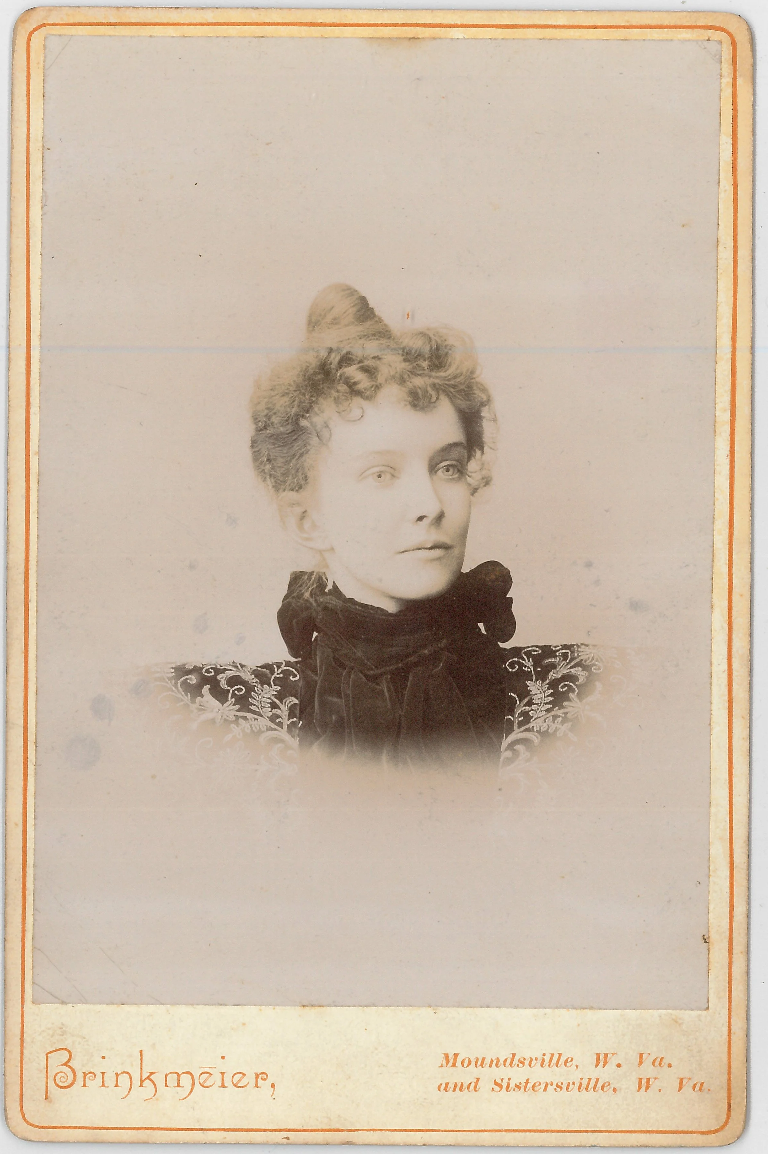 Vintage sepia-toned portrait photograph of a woman with curly hair styled in an updo, wearing a dark dress with intricate embroidery and a large bow or scarf around her neck, mounted on a card that reads 'Brinkmeier, Moundsville, W. Va. and Sistersville, W. Va.' in the bottom corner.