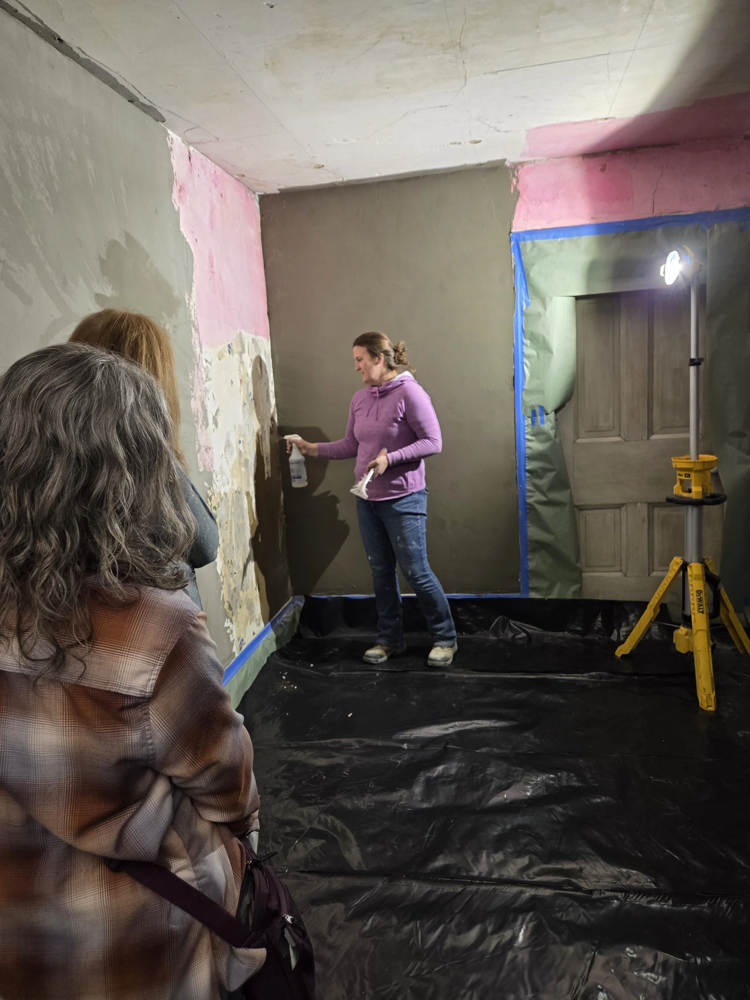 A woman demonstrating plaster repair  techniques to a group inside a room under construction, with plastic sheeting on the floor and partially painted walls.