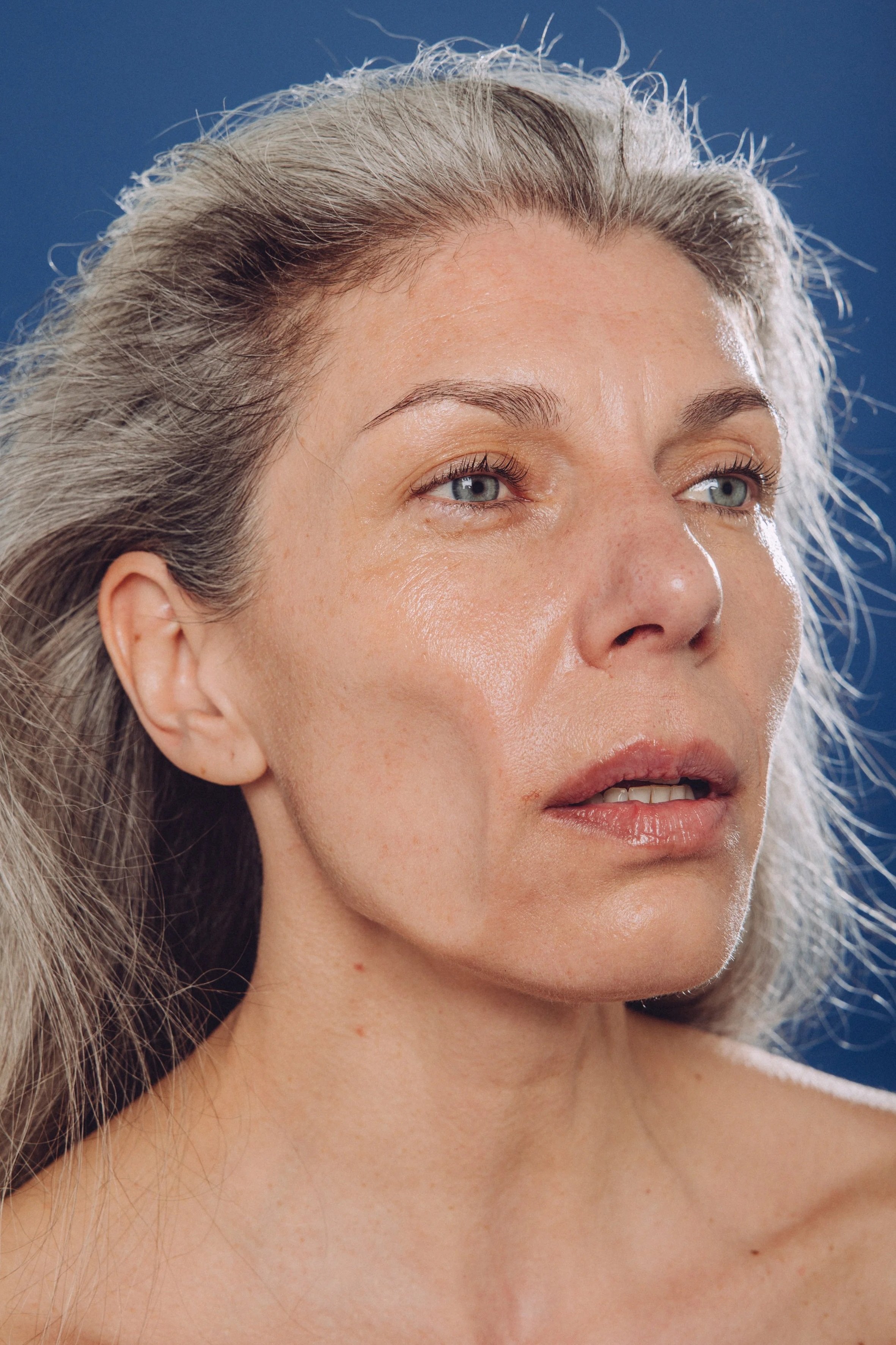 A close-up portrait of an older woman with long, wavy gray hair, against a dark blue background. Her cheek is blown around via high speed wind.