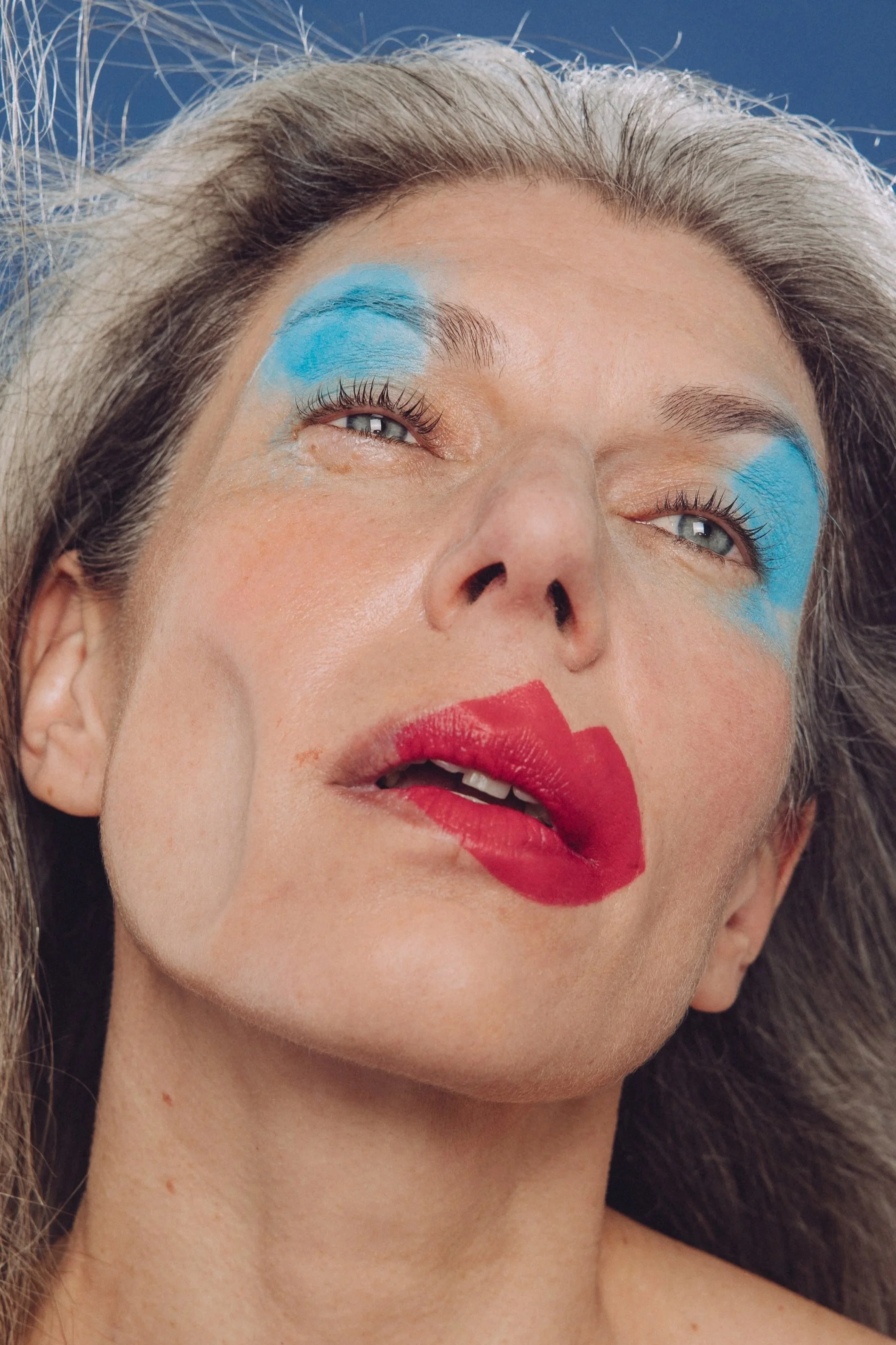 An older woman featuring blue eyeshadow and scarlet lipstick that looks out of place, as if the wind has blown her makeup off into the right hand corner of the screen, her cheek is also blown inwards as if by high pressure wind.