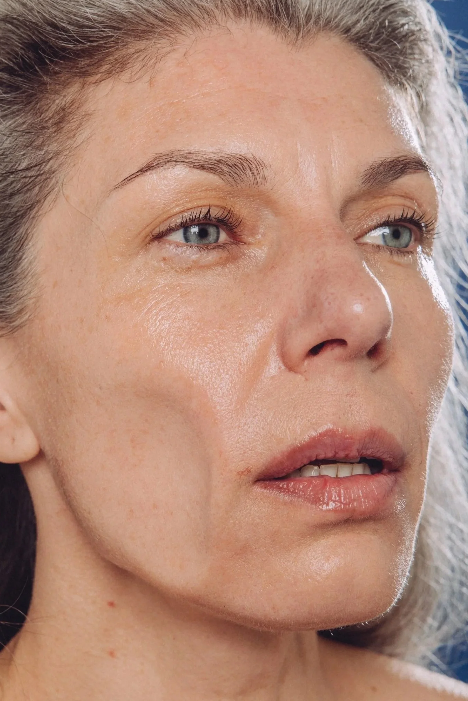 A close-up portrait of an older woman with long, wavy gray hair, against a dark blue background. Her cheek is blown around via high speed wind.