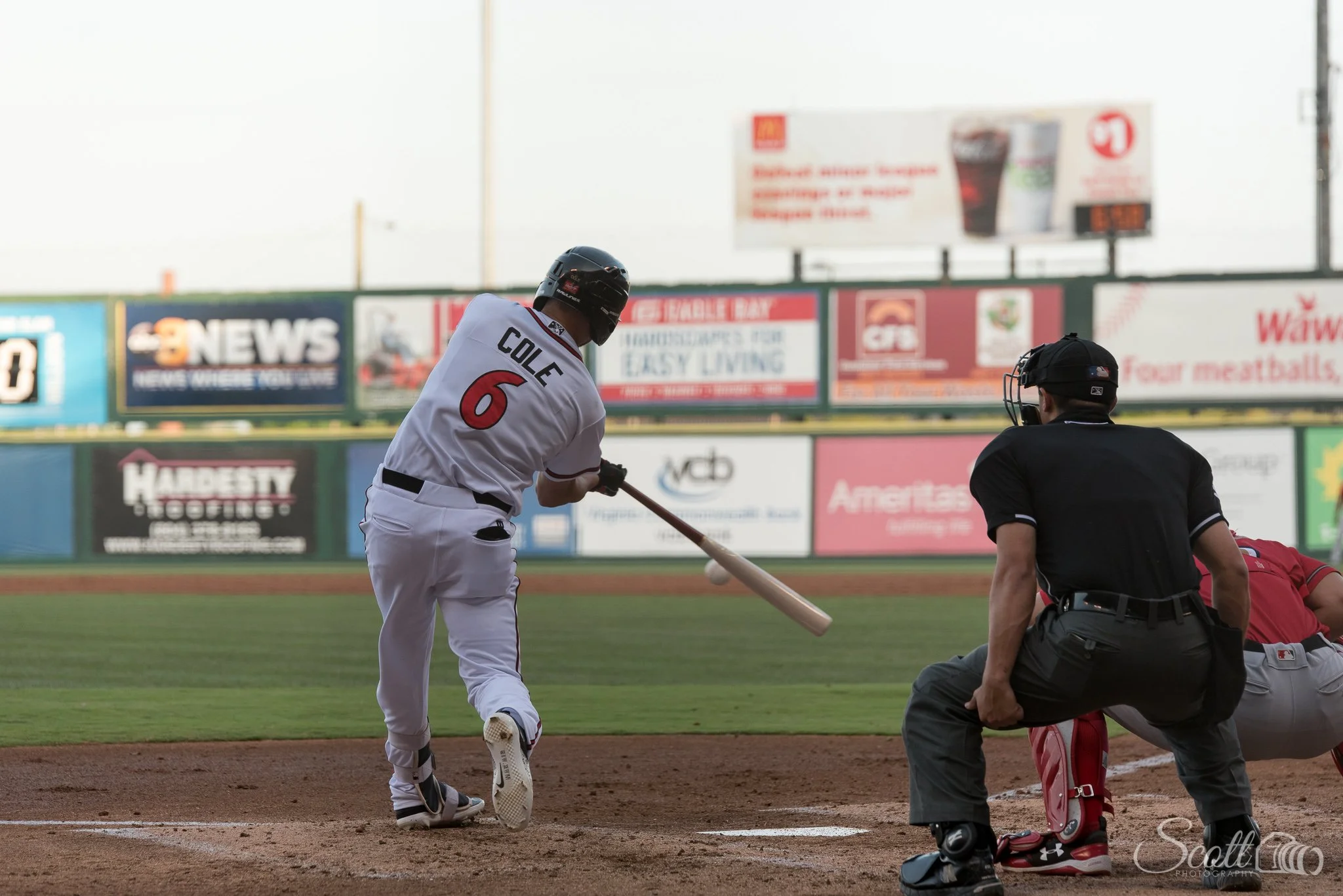 Baseball player making contact at the plate during a live game, captured from behind the batter by Scott Photography.