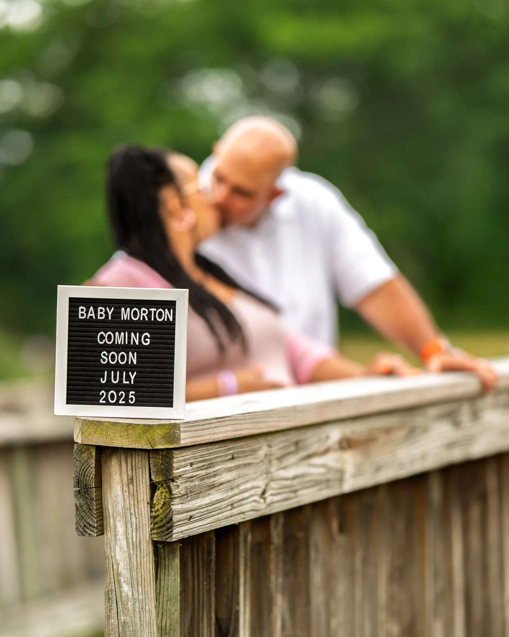 Creative maternity pregnancy announcement photo of a couple on a wooden bridge with baby sign, photographed outdoors in Central Virginia by Scott Photography.
