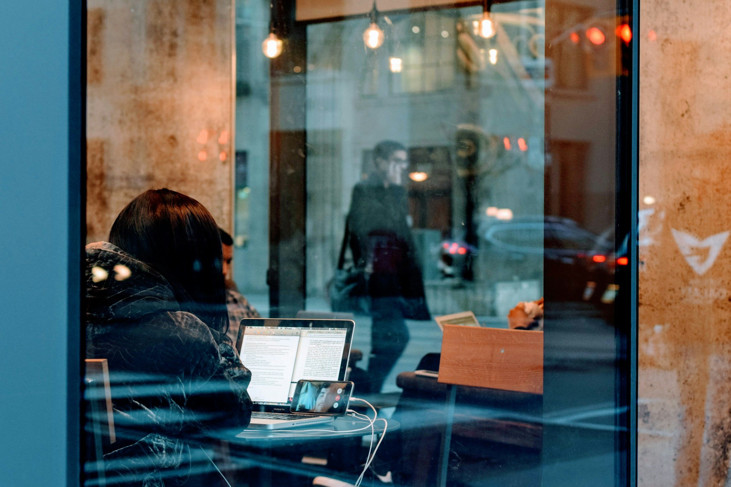 Café sitzt Person mit Laptop, in der Scheibe spiegelt sich ein Mann