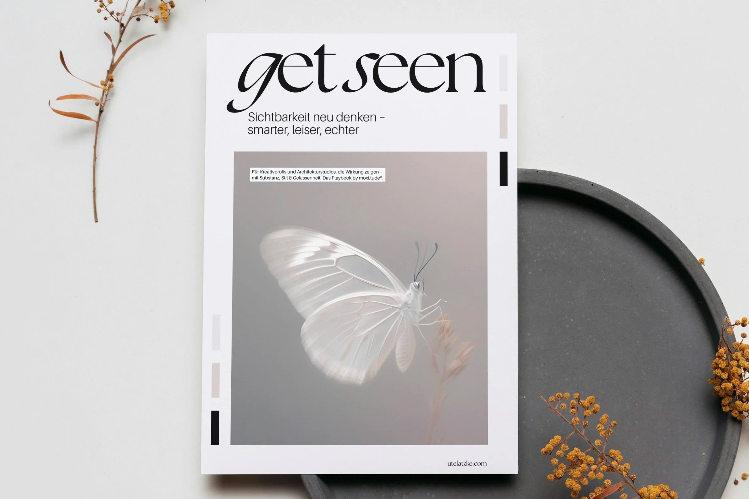 Cover von GetSeen mit großem Schmetterling liegt auf grauem Teller, auf Schrebtisch