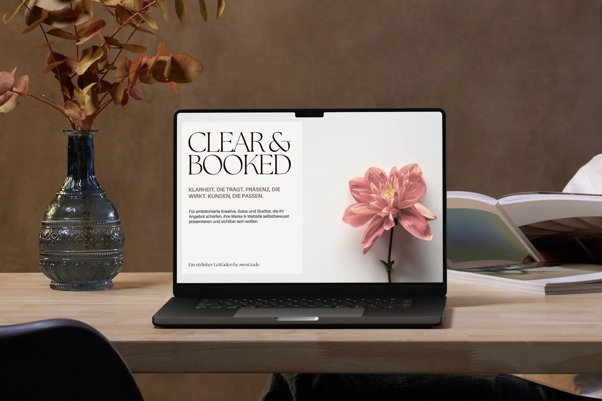 Macbook auf Schreibtisch vor brauner Wand, daneben links Glasvase, zeigt Cover von Clear&Booked