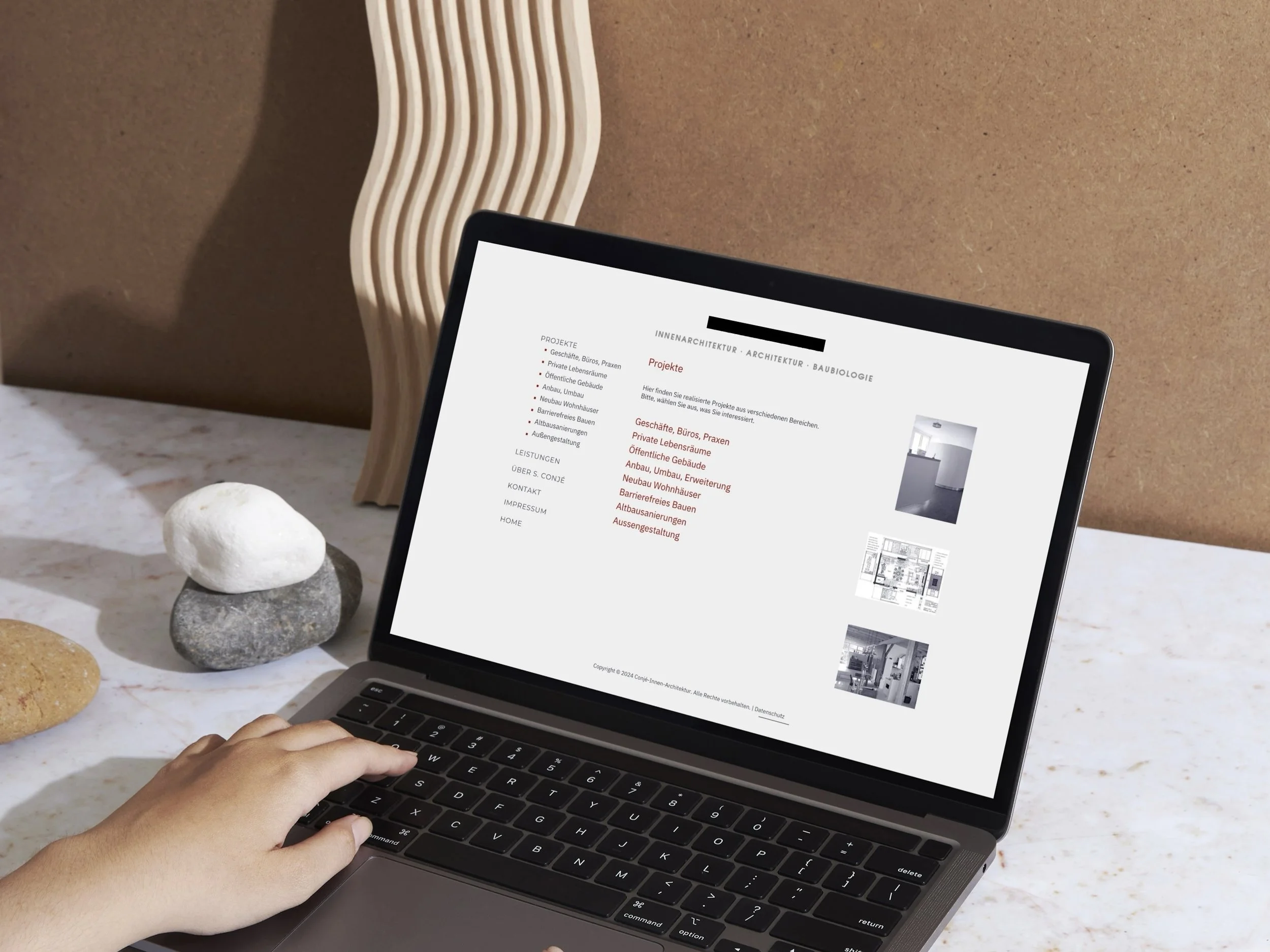 Macbook auf Tisch vor brauner Wand, zwei Steine und eine Hand