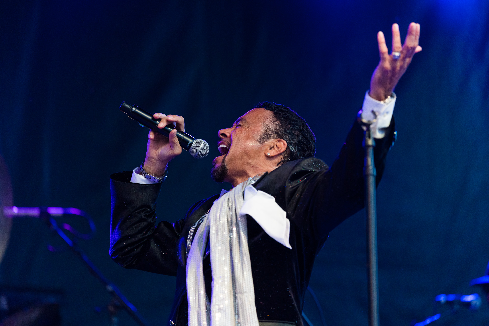 Morris Day and the Time passionately performing on stage, holding a microphone close to his mouth with one hand raised in the air.