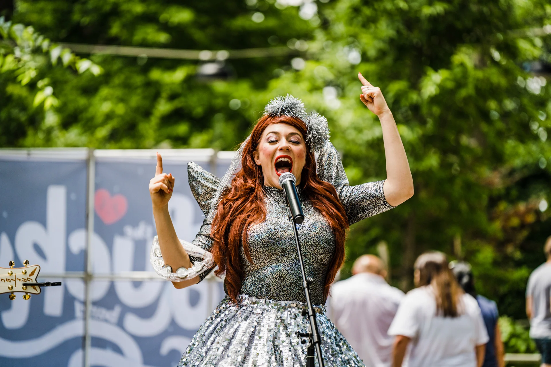 Alisha Gaddis of Lucky Diaz and the Family Jam Band wearing a silver sequined dress and tinsel headbands singing into a microphone at an outdoor event with a crowd and trees in the background.