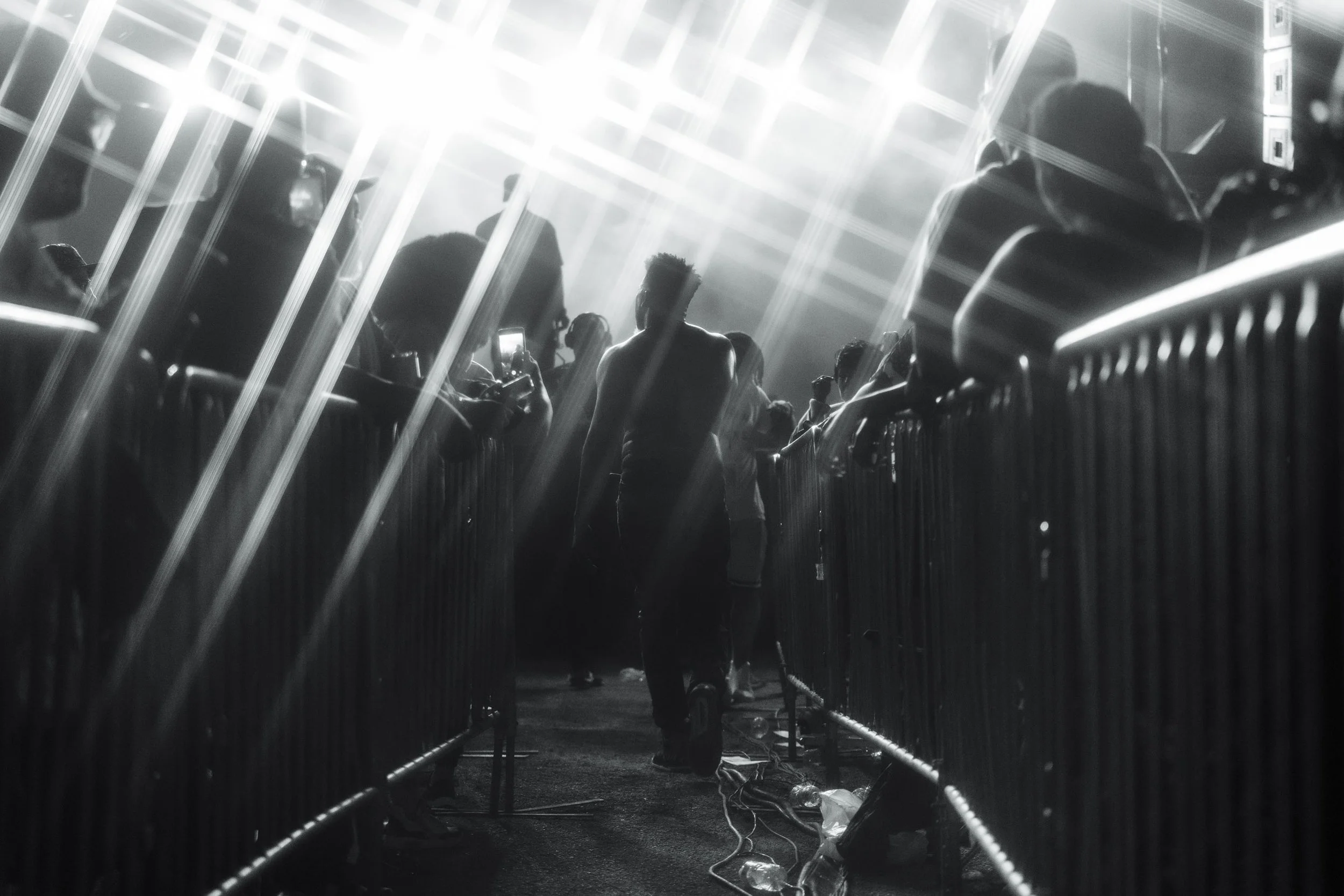 Crowd of people at a David Banner concert with bright lights from above illuminating the scene. People are standing behind a waist-high barrier, some taking photos or videos with their phones. The photo is in black and white.