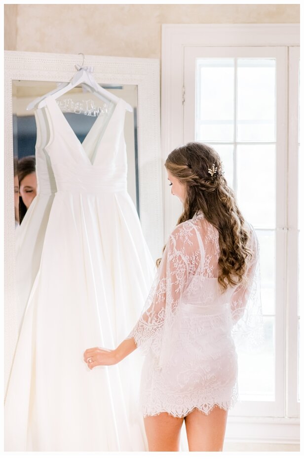 bride wearing a white robe looking at her wedding dress in mirror