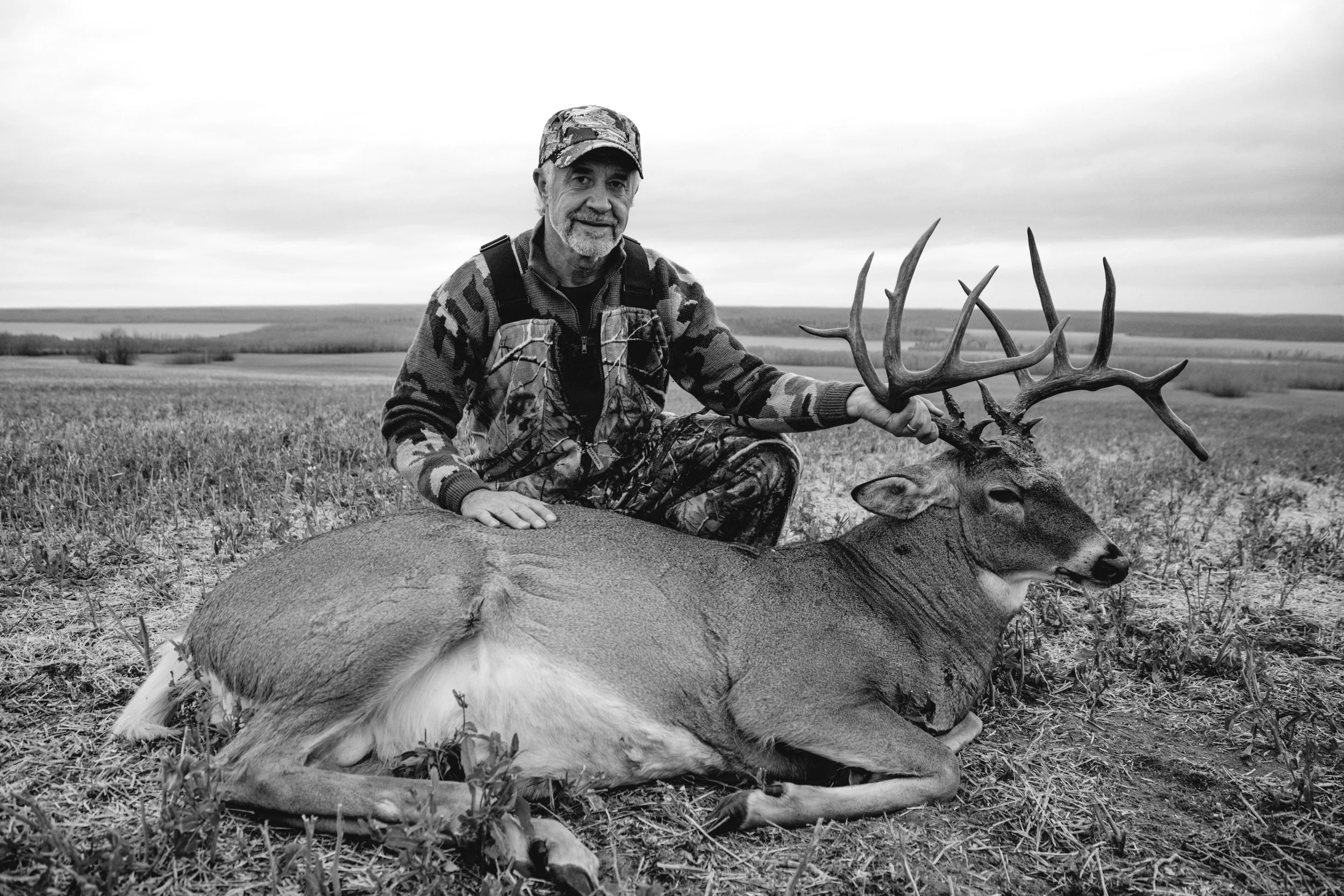 A man in camouflage hunting gear kneels next to a large deer with antlers, holding its antlers in an open field.