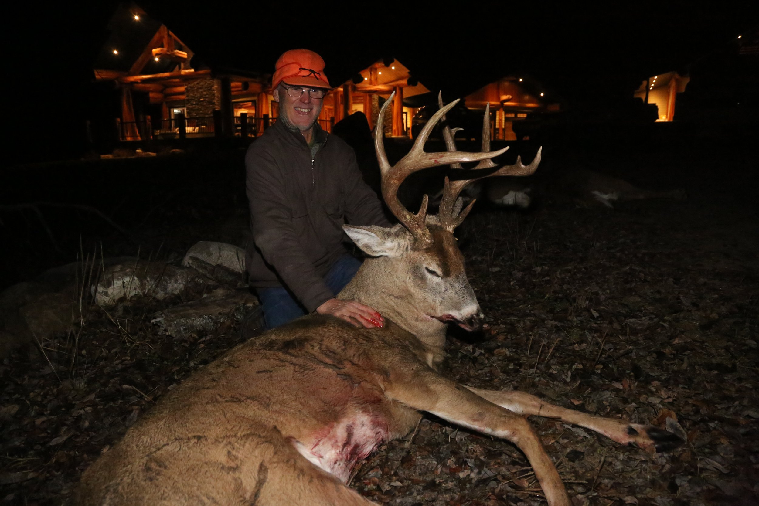 Man kneeling next to a large, dead deer with antlers, during nighttime with lit log cabin in background