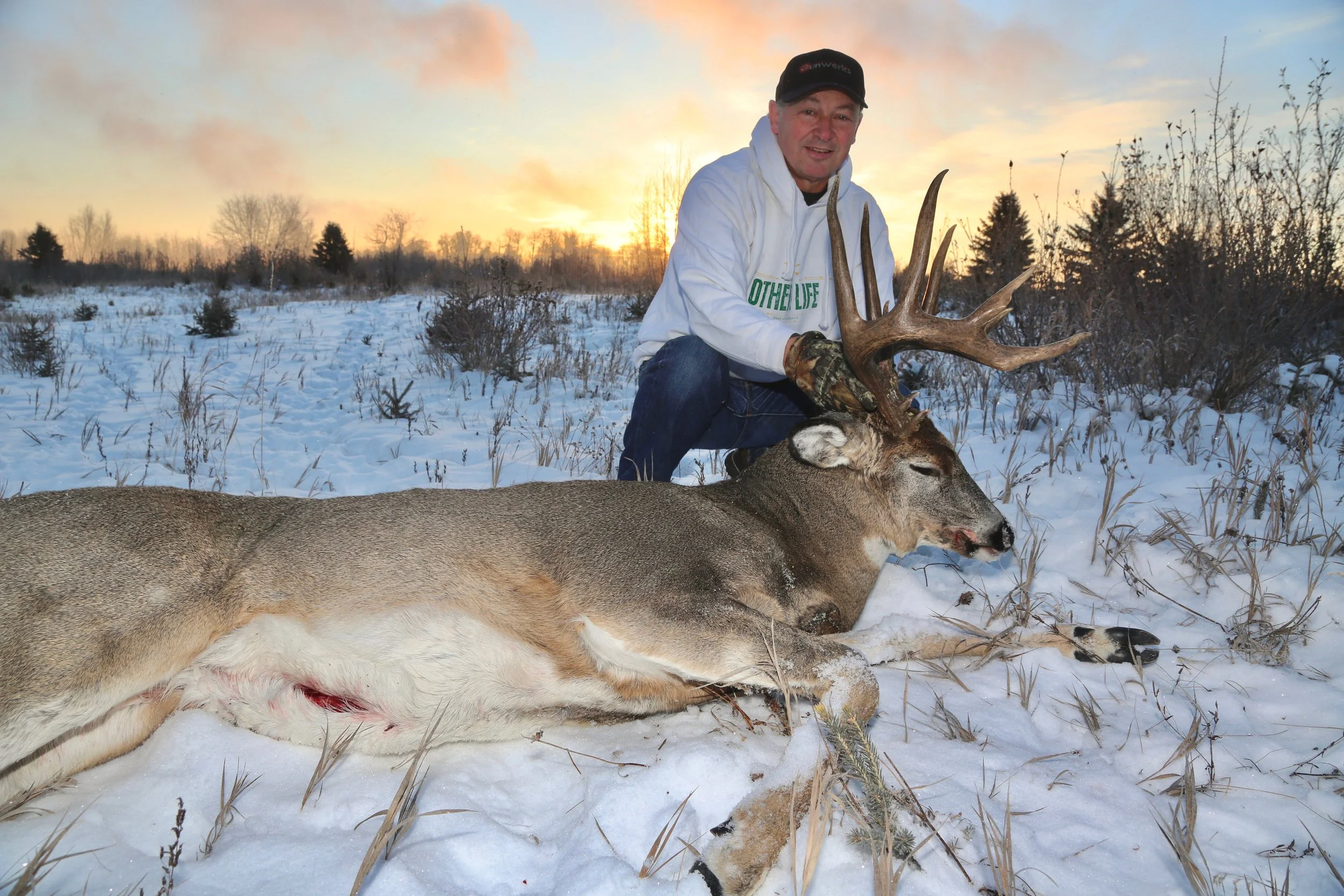 A man in winter clothing kneels on snow-covered ground, holding the antlers of a large, dead deer with a full set of antlers. The deer is lying on its side, and the background features a sunset sky with trees and sparse winter vegetation.