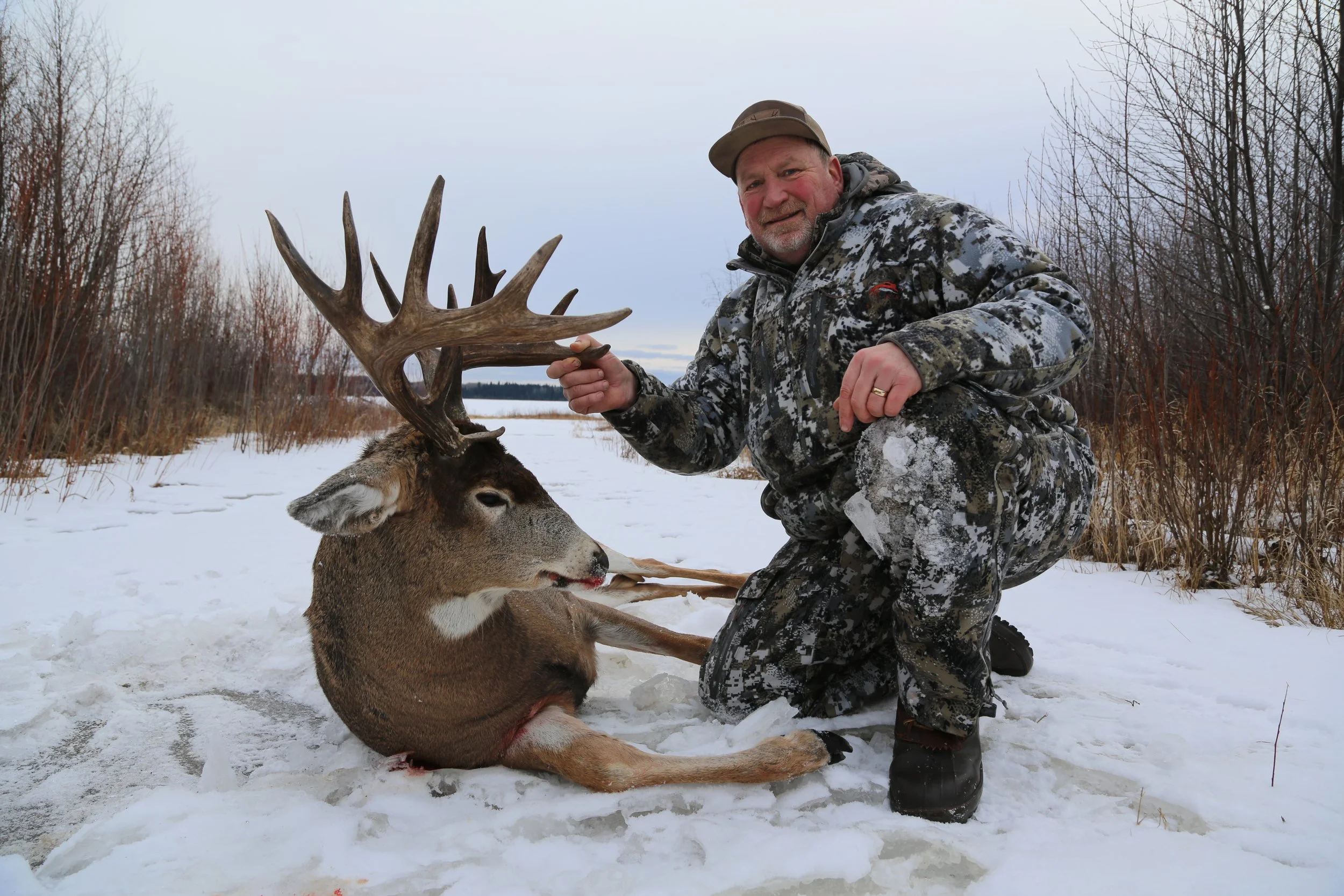 A man dressed in camouflage clothing kneels in the snow beside a freshly hunted deer with large antlers, holding the antlers and smiling at the camera on a snowy landscape with leafless trees in the background.