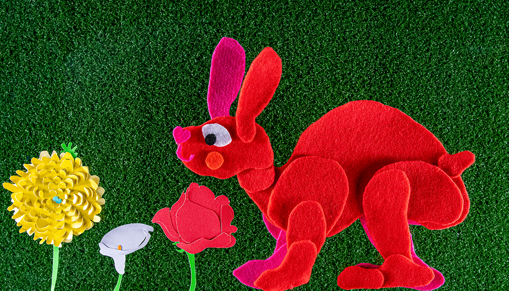 A red plush rabbit sitting on green grass next to yellow, white, and red paper flowers.