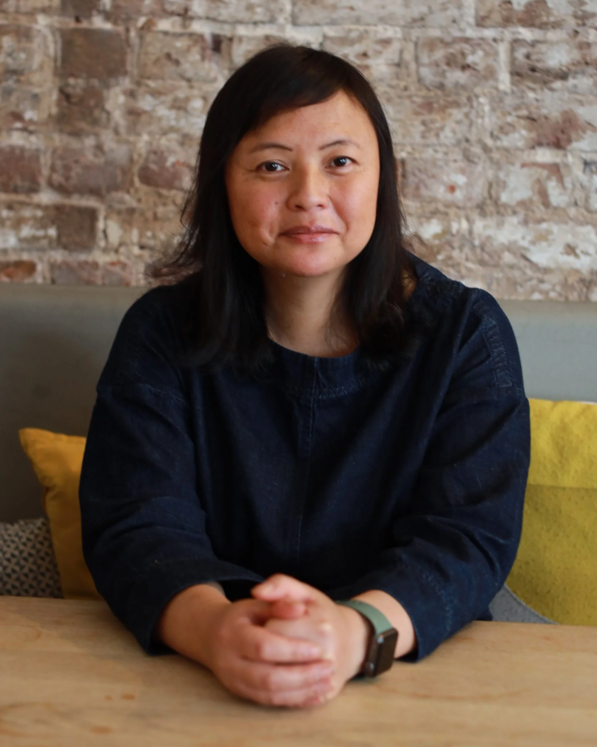 A woman with dark hair and Asian features sits at a wooden table in front of a brick wall, wearing a dark denim dress and a black watch, with her hands clasped together on the table.