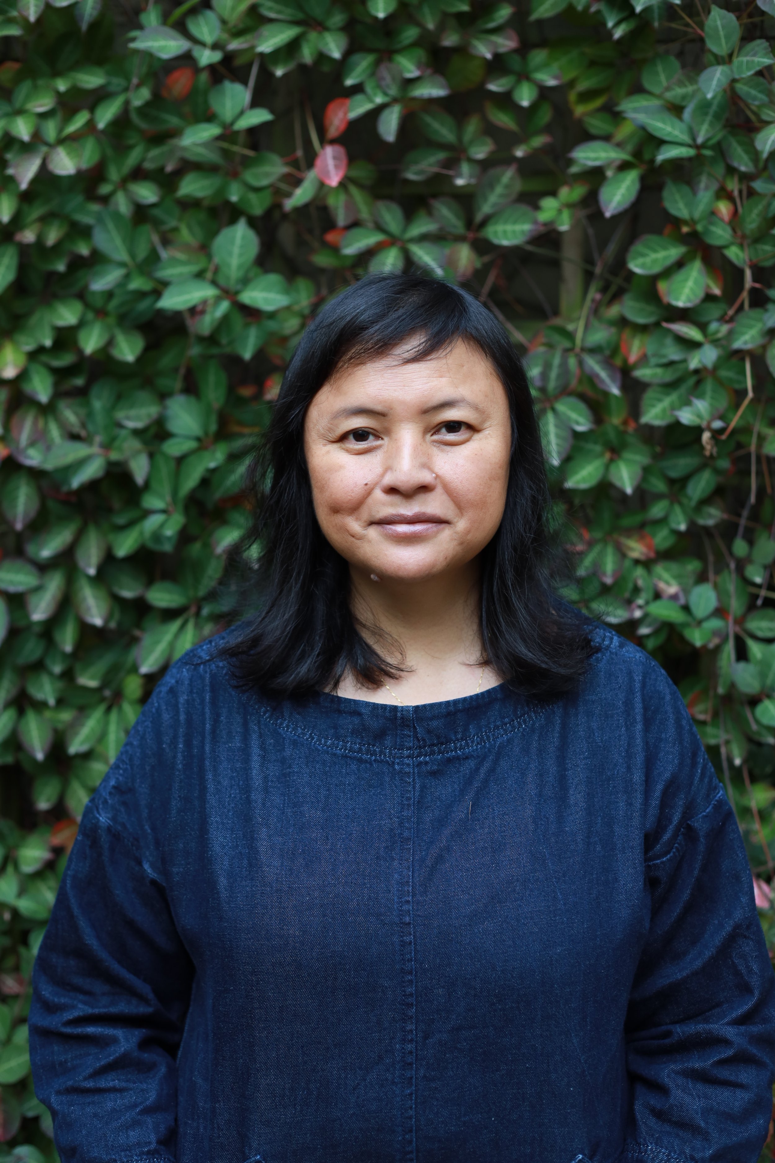 Portrait of a woman standing outdoors in front of green leafy plants, wearing a dark blue denim dress.