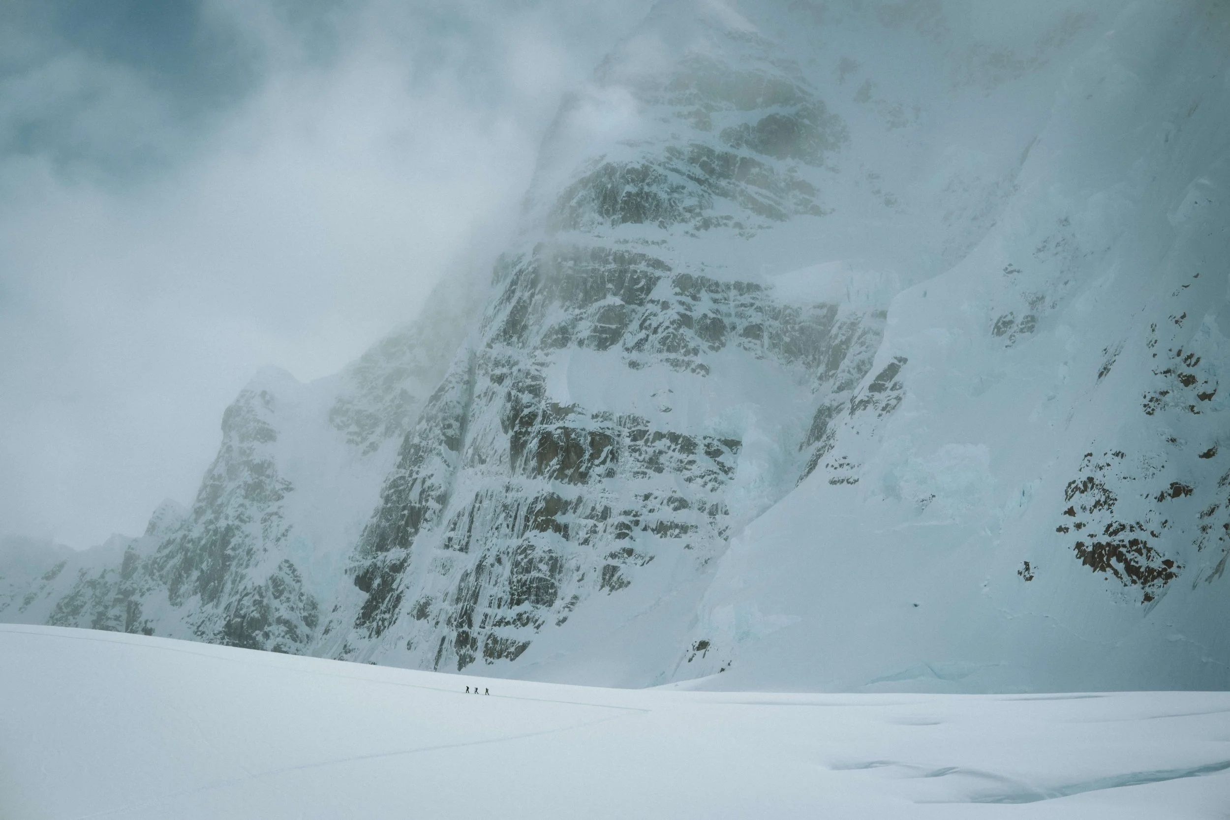three climbers ski towards the base of The North Buttress of Mount Hunter, Alaska, which looms above them in the snow.