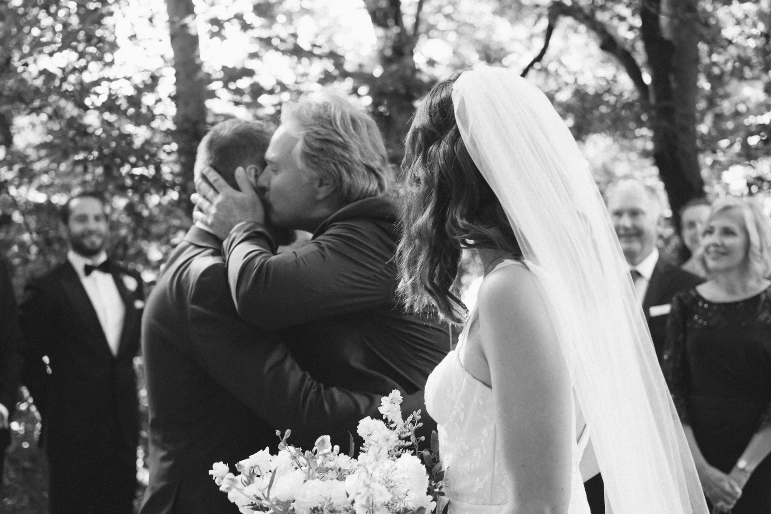 Groom hugging his father during an emotional wedding ceremony as the bride looks on