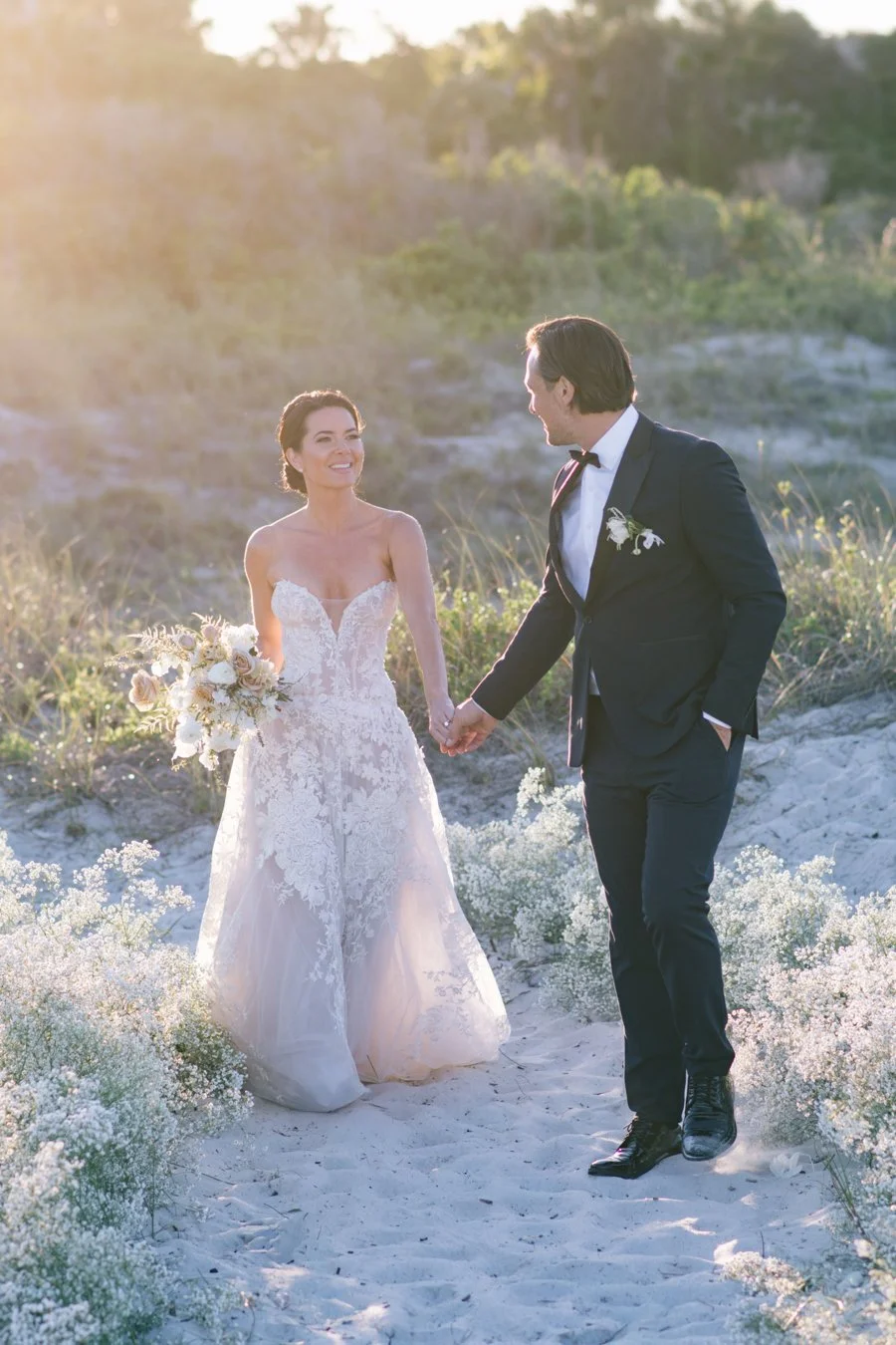 Bride and groom walking hand in hand along a sandy path during their destination wedding, surrounded by soft greenery and natural light.