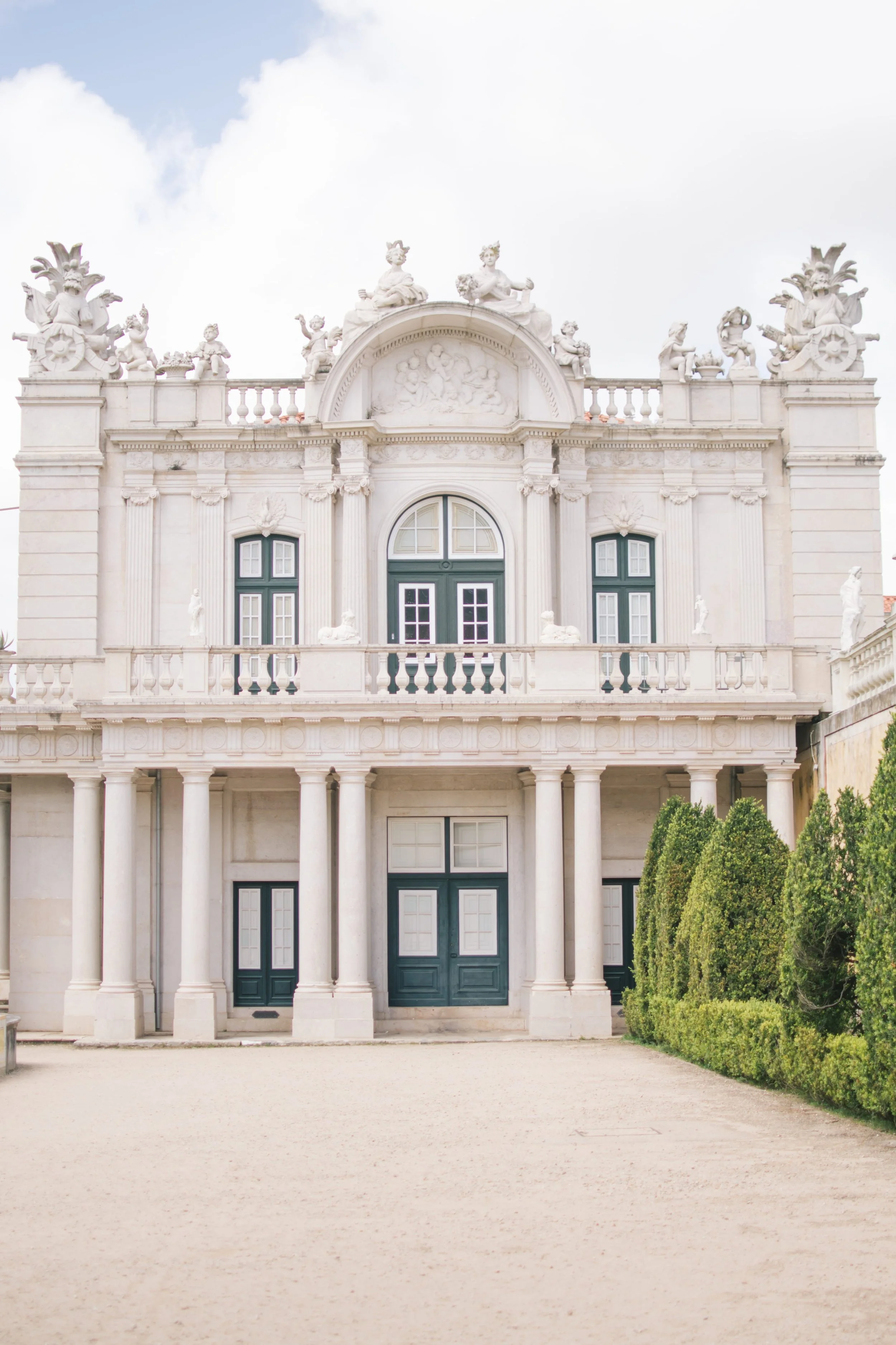 Exterior view of a grand white neoclassical palace wedding venue in Portugal, Palácio de Queluz, featuring ornate stone sculptures, a grand balcony, and manicured gardens.