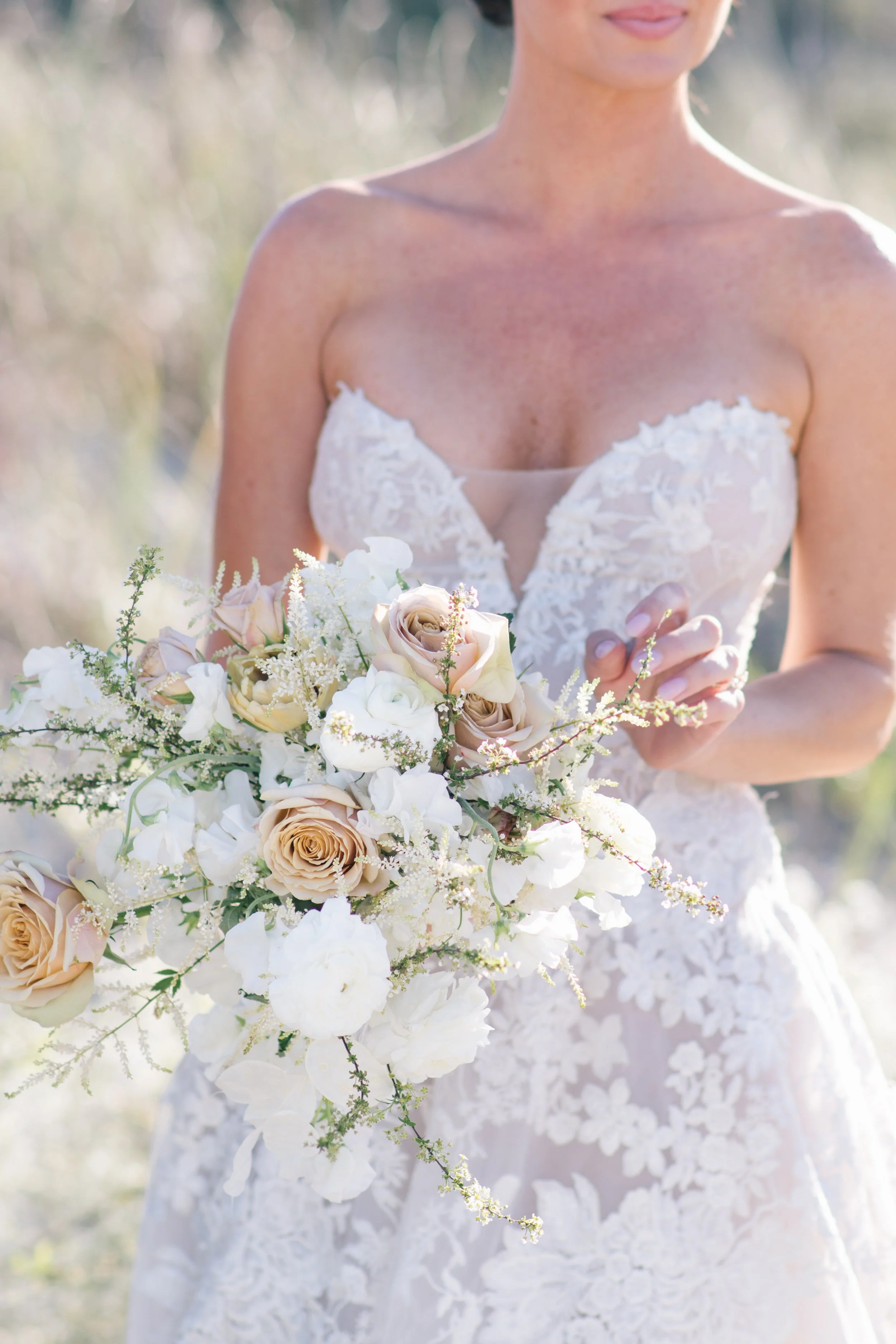 Bride holding a soft neutral floral bouquet during her destination wedding, wearing a lace wedding gown in natural light.