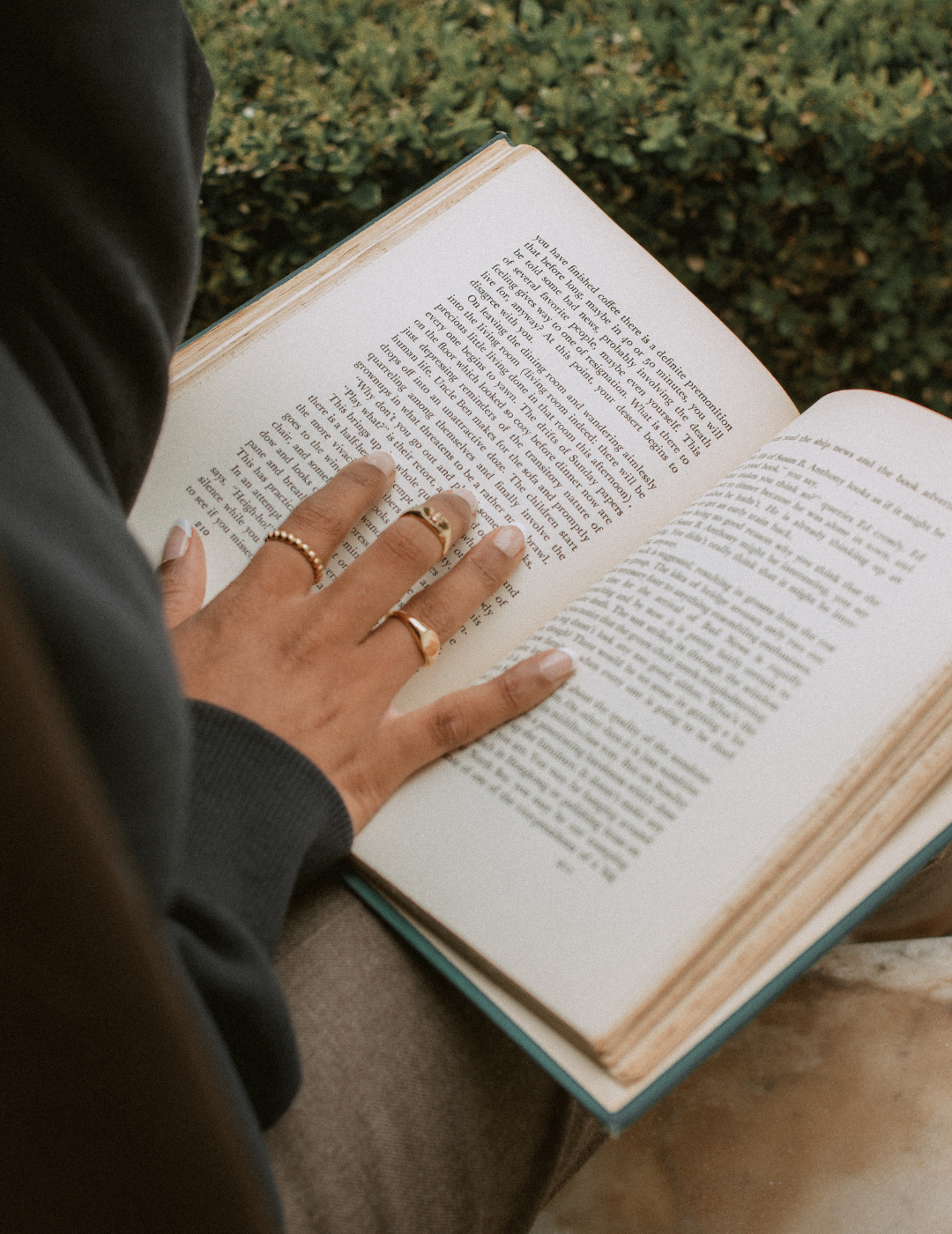 Person reading a book outdoors, wearing rings and a dark jacket, with text visible on the pages.