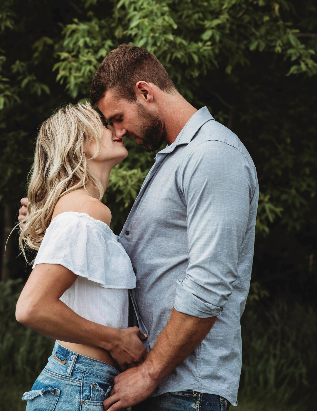 A man and woman about to kiss outdoors with greenery in the background.