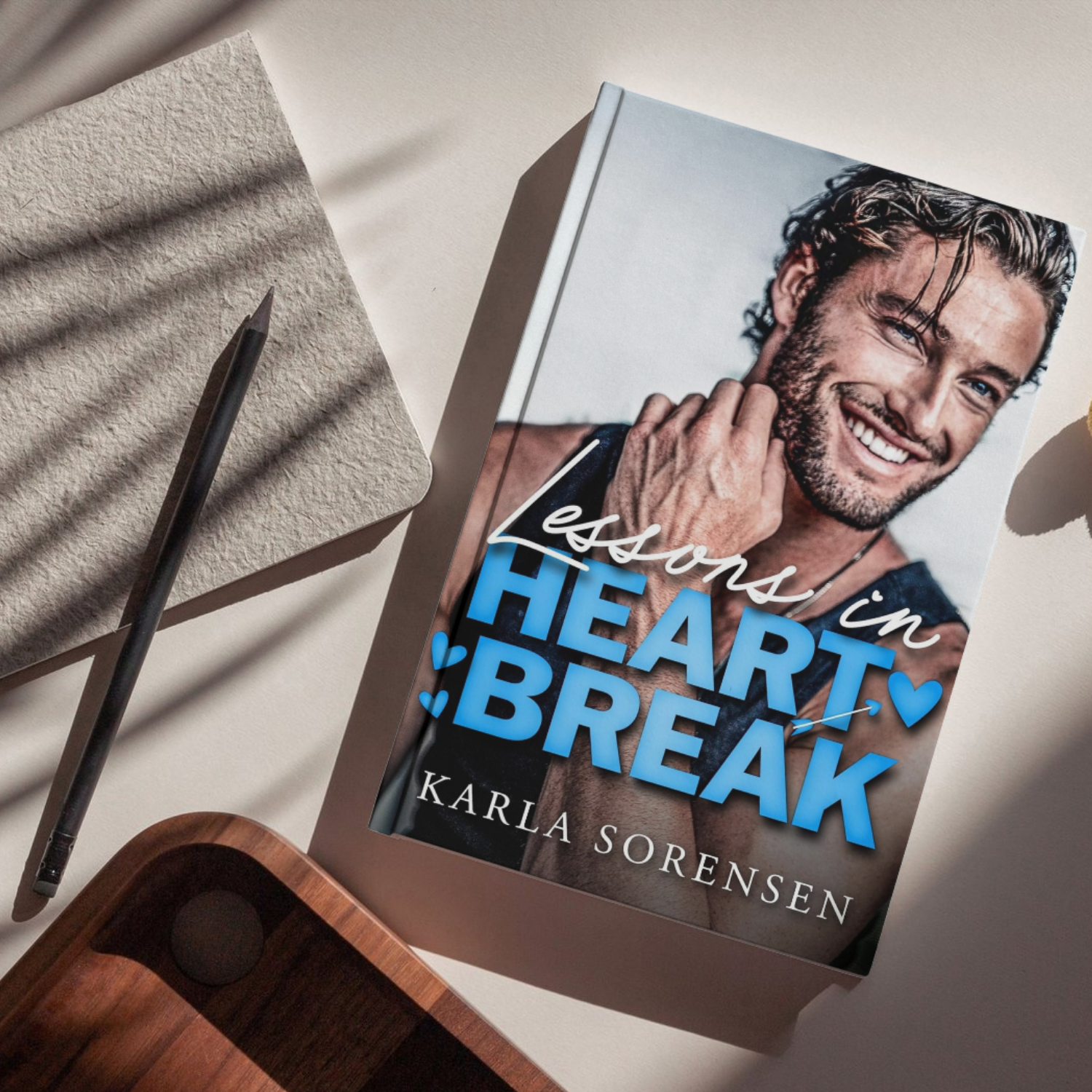 Book titled 'Lessons in Heartbreak' by Karla Sorensen, featuring a smiling man with wavy hair and blue eyes on the cover, with a beige textured notebook, a black pen, and a wooden tray nearby on a white surface.