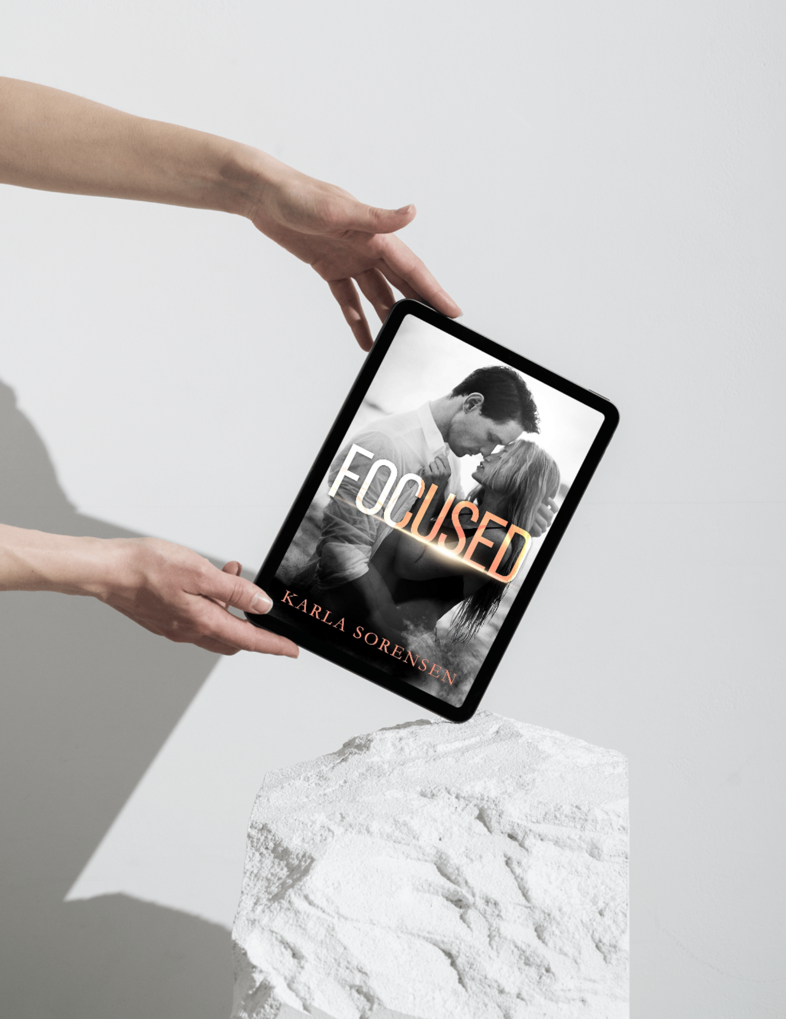 A person holding a tablet displaying a book cover titled 'FOCUSED' by Karla Sorensen, with a black-and-white photo of a man and woman close together, in front of a white textured surface.