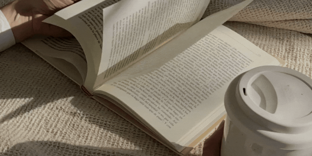 A person reading a book while holding a disposable coffee cup, resting on a textured surface with natural light.