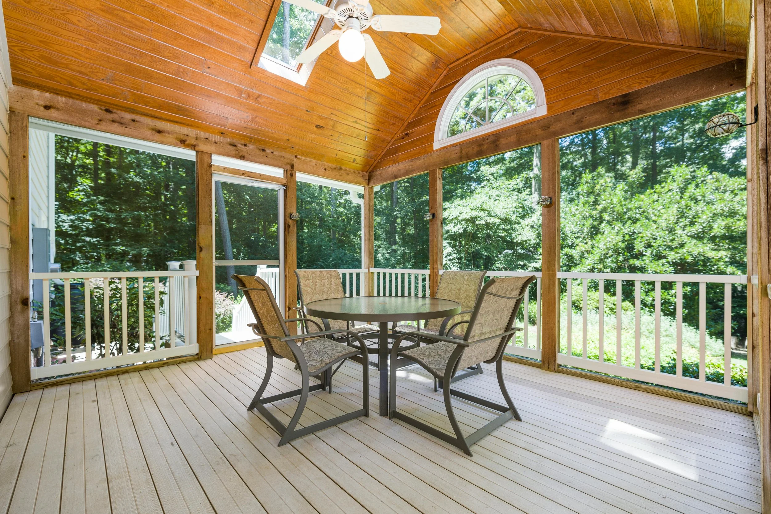 Screened wooden porch with a vaulted wood ceiling, ceiling fan, round outdoor table with four chairs, and forest views through large screened panels.