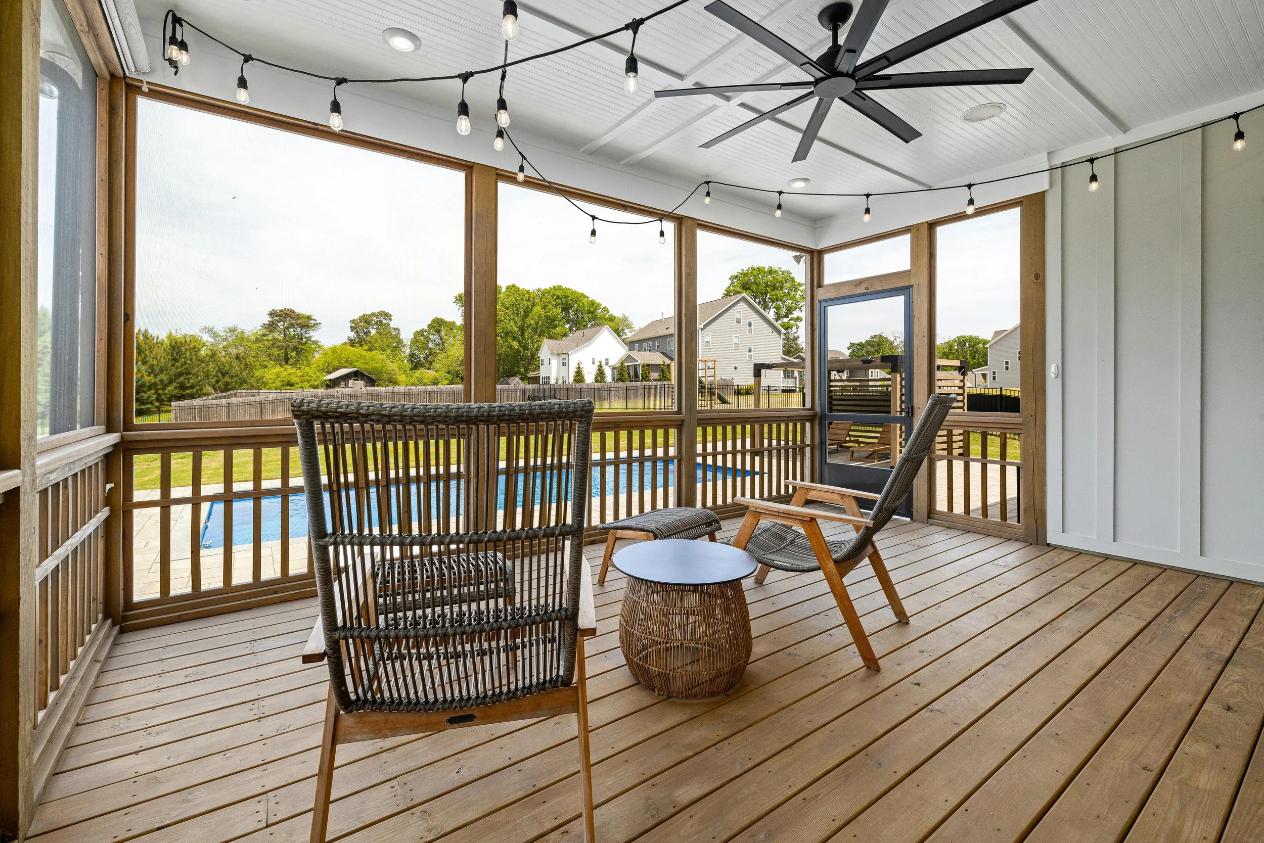 Modern screened porch with wood decking, woven lounge chairs, a small accent table, ceiling fan, and string lights, overlooking a backyard pool and nearby homes.