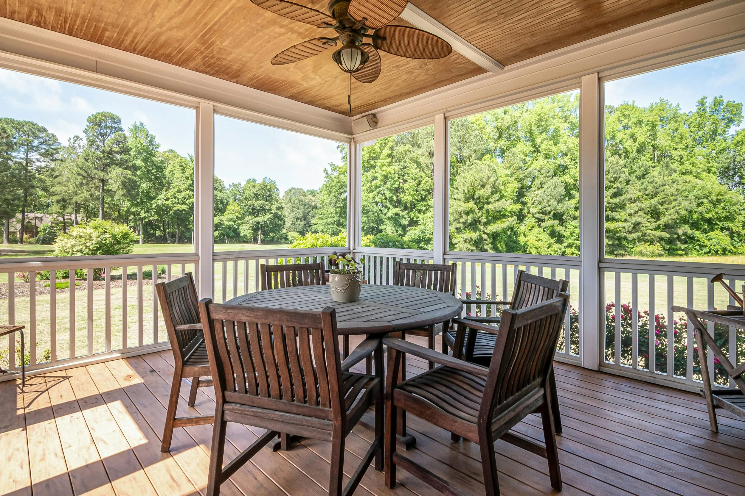Covered screened porch with a wooden dining table and chairs, ceiling fan overhead, and open views of a green backyard and trees through white railing.