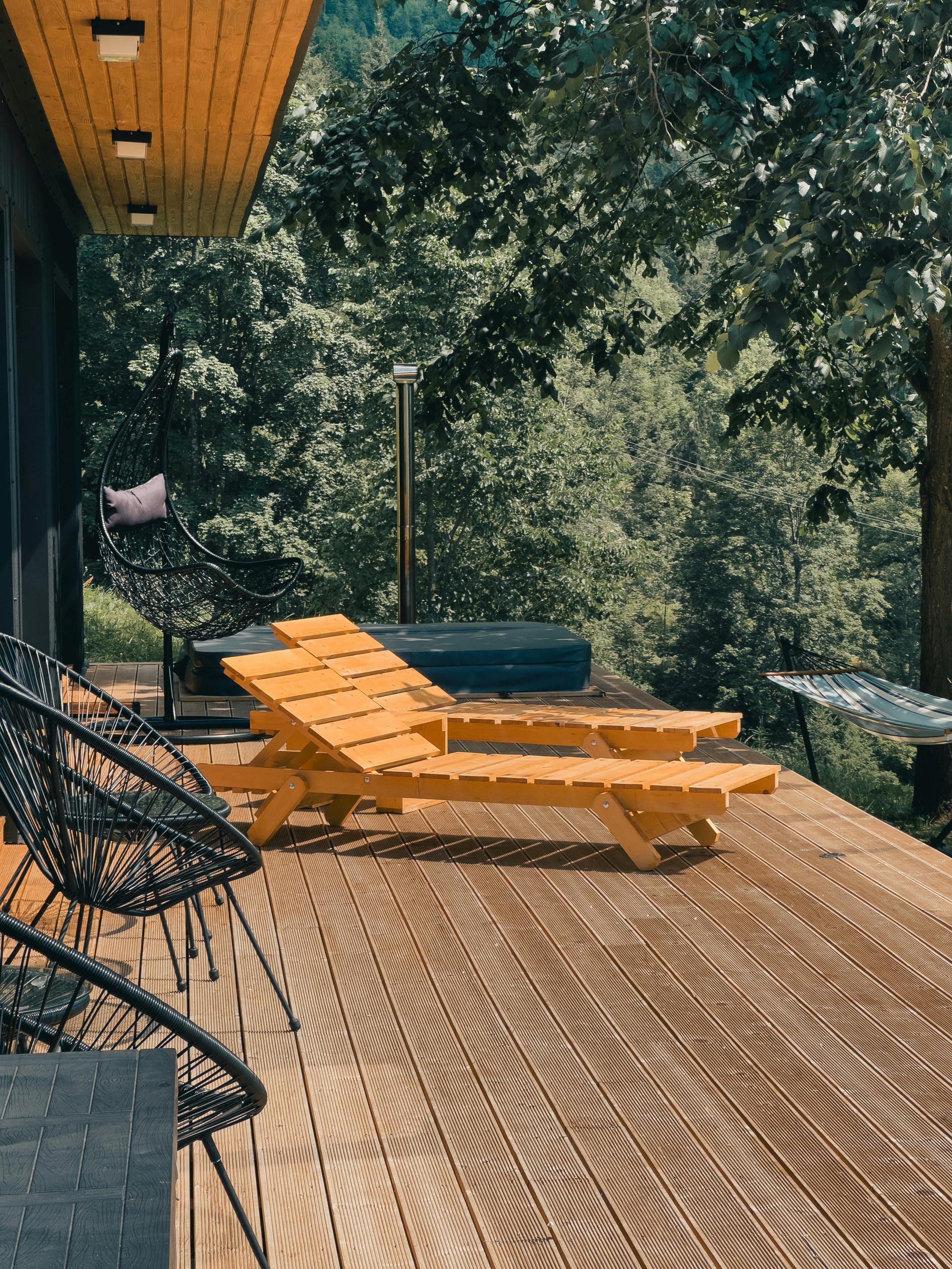 Wooden deck with a picnic table and built-in benches on a modern cabin patio, surrounded by trees, featuring black outdoor chairs, a hanging hammock chair, and forest views in the background.