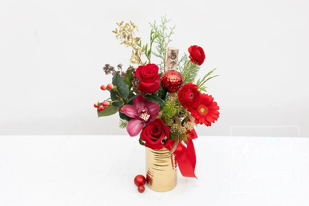 A winter holiday arrangment in a tall gold vase featuring red flowers , evergreens, and gold accents.