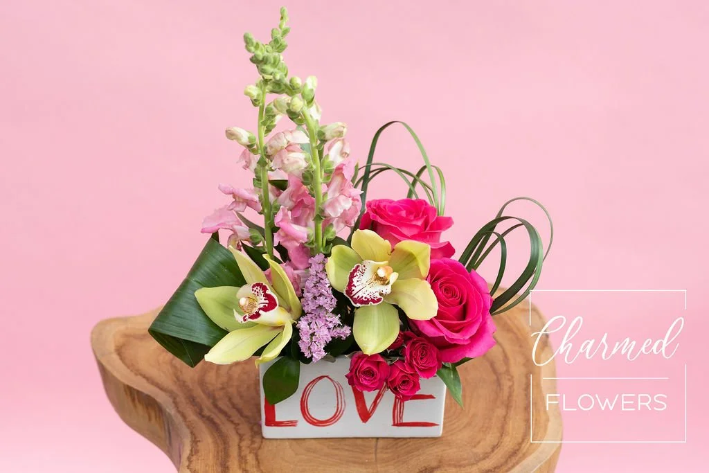 The Love Vase is a long and low vase with the word Love written on it. It is a colourful bright arrangment with orchids and roses, perfect for your valentine