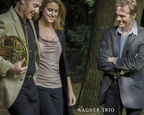 Three people standing outdoors in front of trees, smiling and engaging with each other. One person holds a French horn. The text "Wagner Trio" is at the bottom.