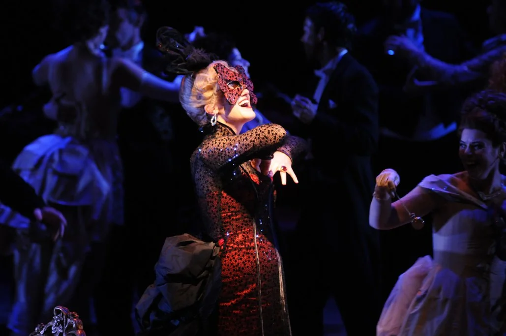 A woman in a Venetian-style mask and ornate dress dances and laughs among other costumed performers on stage.